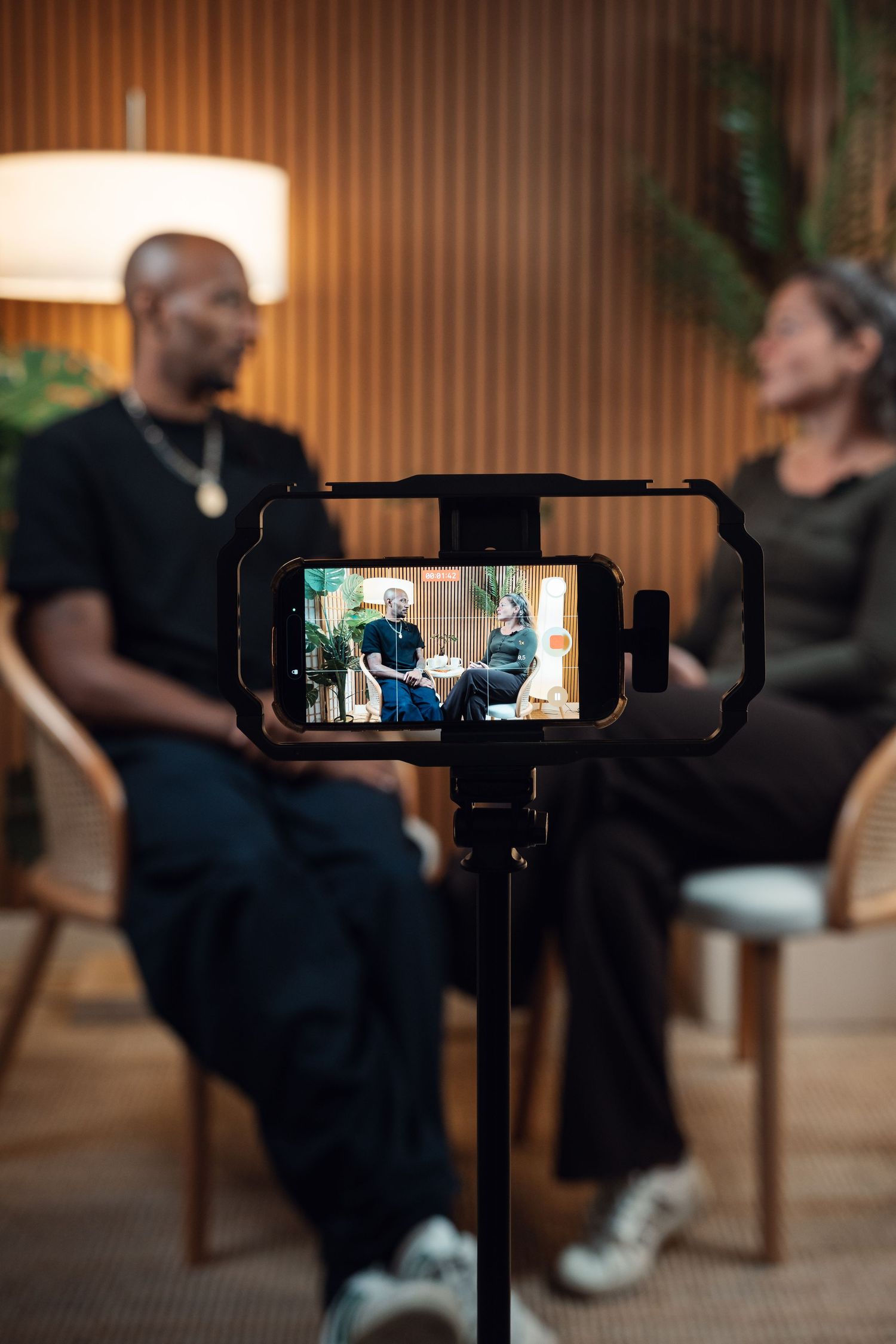 Two people being filmed on a smartphone mounted on a tripod during an interview session in a warmly lit studio.