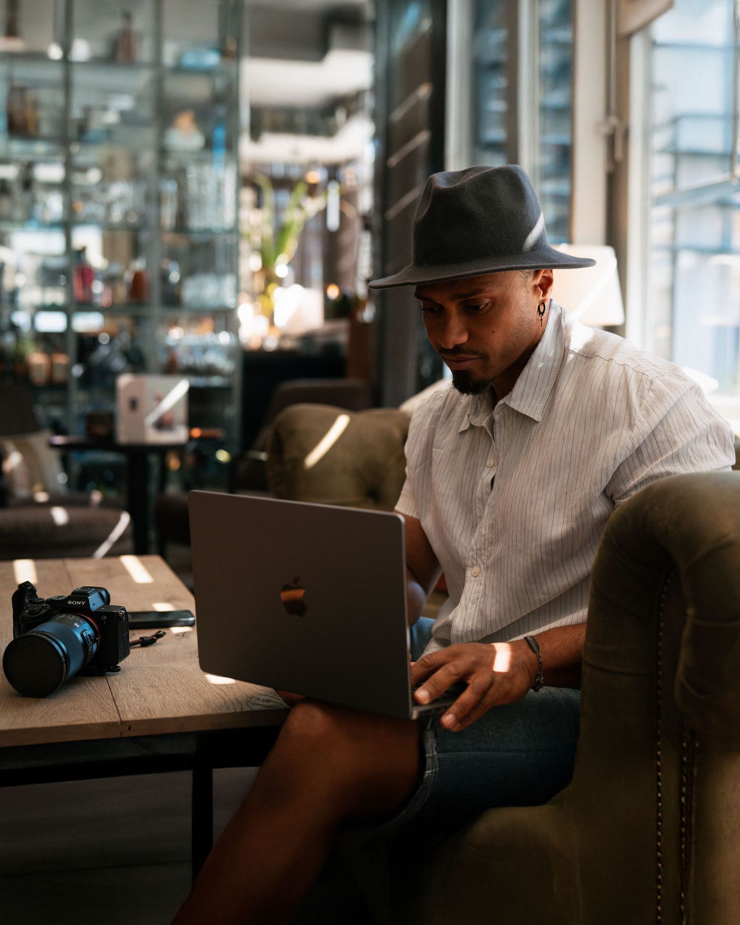 Man in gray fedora hat working on laptop in stylish cafe with camera on table beside him.