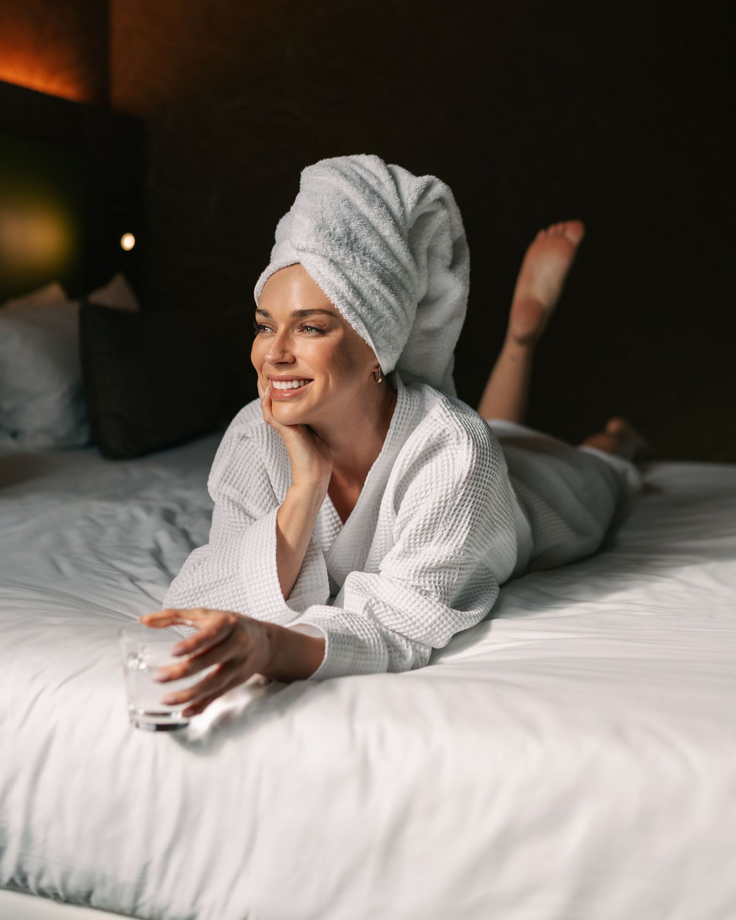 Relaxed woman in white bathrobe lying on hotel bed holding glass of water.