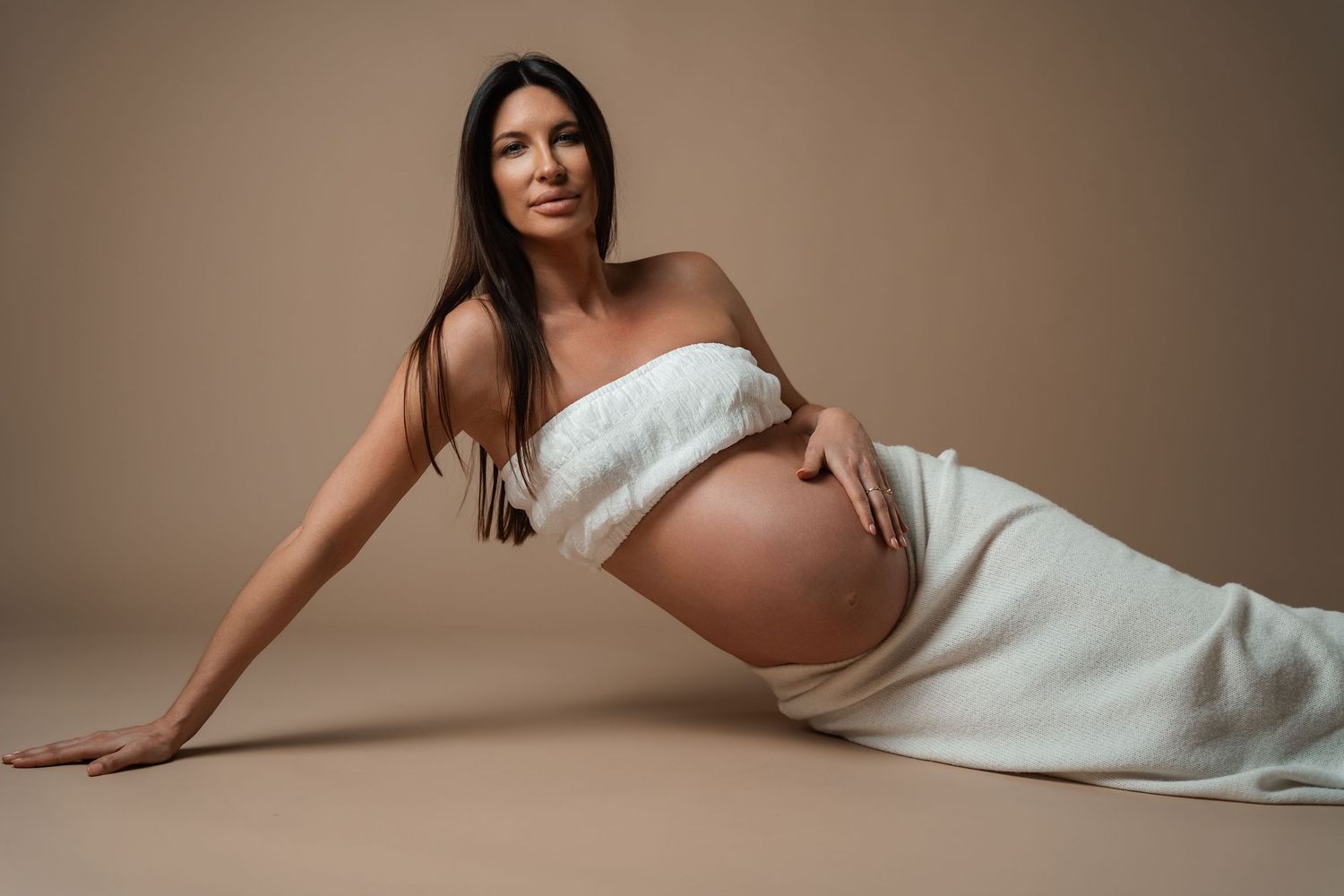 Pregnant woman in white wrap poses gracefully on studio floor against beige background, showcasing baby bump.