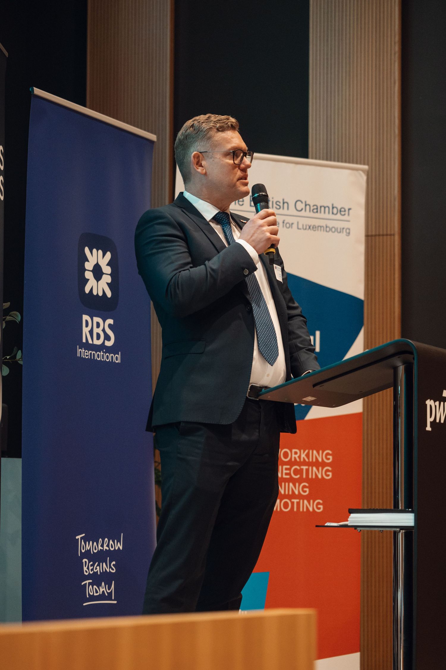 A man in a dark suit speaks at a podium with RBS International and British Chamber Luxembourg banners.