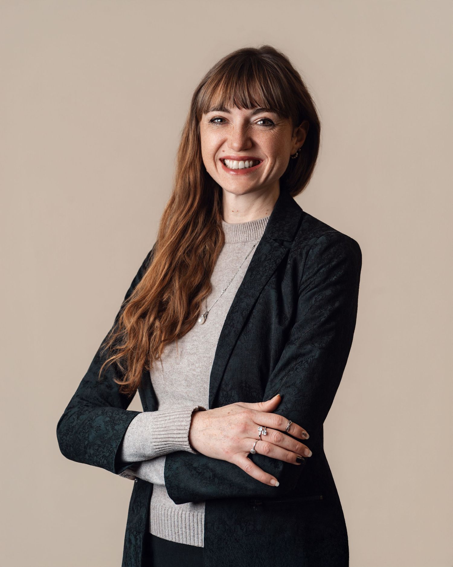 Smiling woman with bangs in a dark blazer and gray turtleneck with arms crossed on beige background.