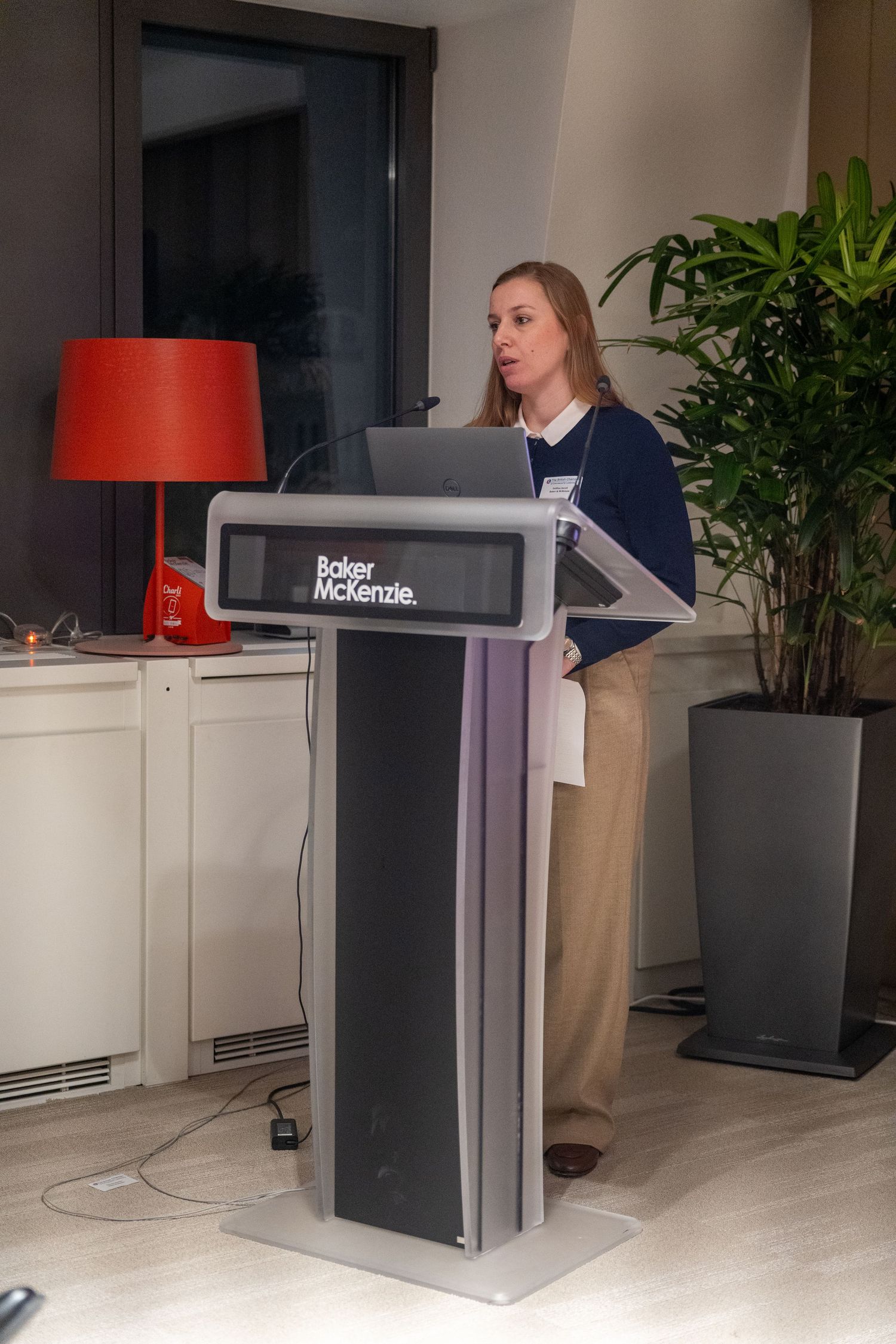 Woman presenting at a Baker McKenzie podium with a laptop at a corporate conference.