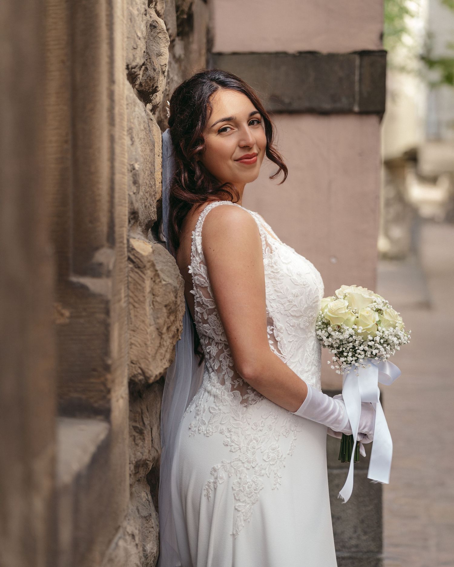 Bride in white lace gown holding bouquet of white flowers, leaning against stone wall in narrow alley.