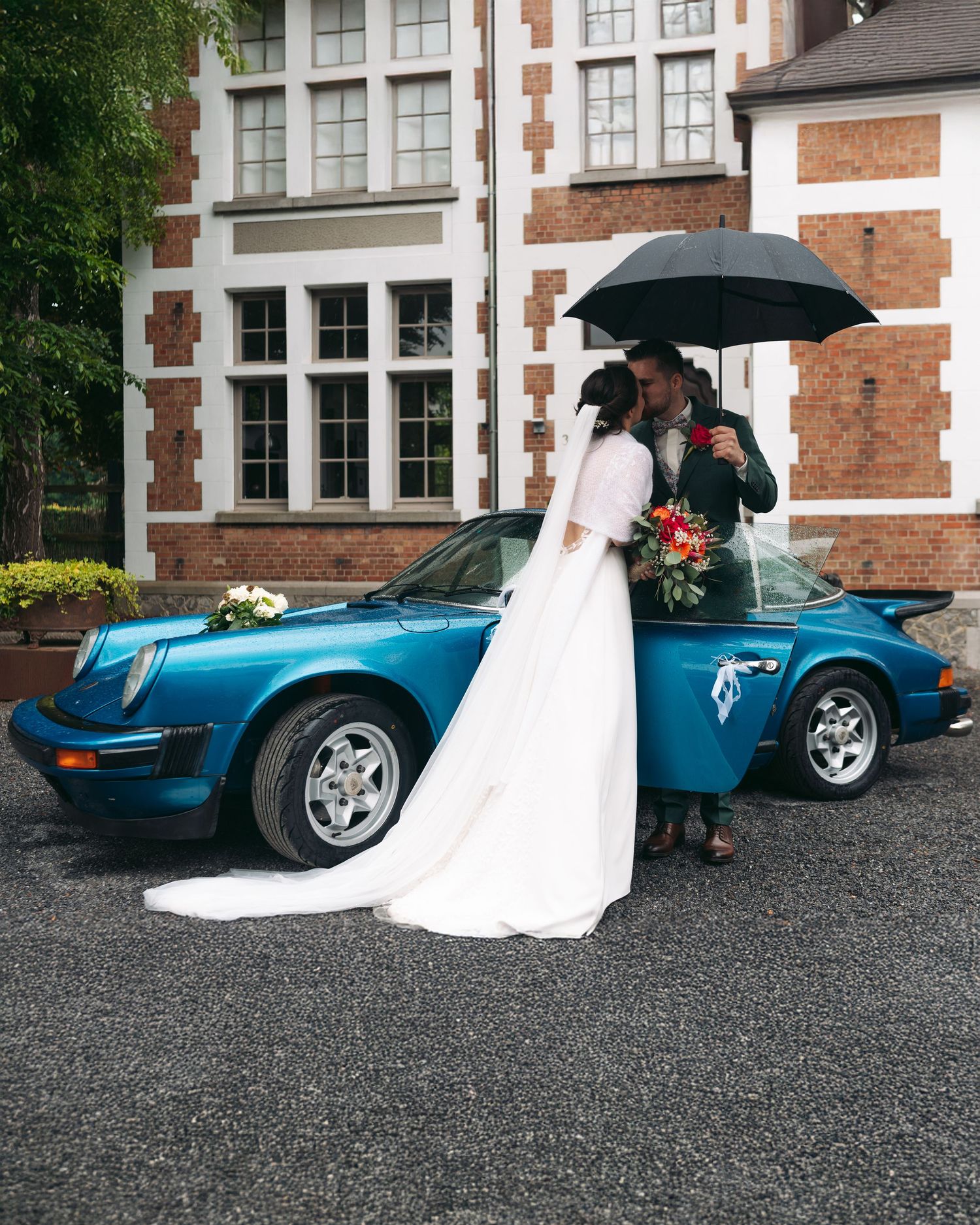 Bride and groom pose kissing beside a vintage blue Porsche 911 under a black umbrella.