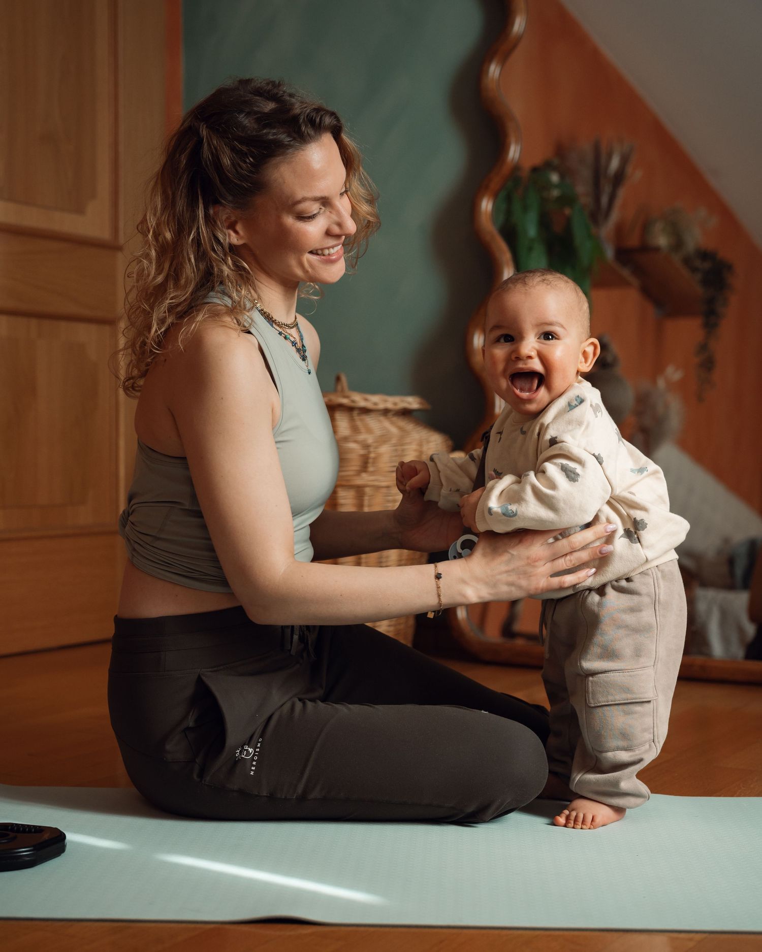 A smiling mother sits on a yoga mat playing with her laughing baby in a cozy home setting.