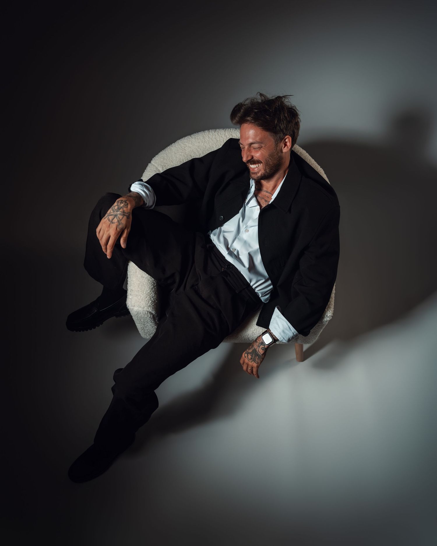 Smiling man with tattoos sits relaxed in a round chair, wearing a black blazer and dark pants in a studio setting.