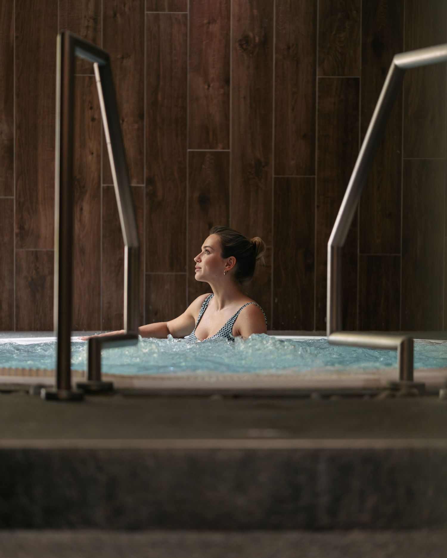 Woman relaxing in bubbling hot tub jacuzzi with wood tile wall backdrop.