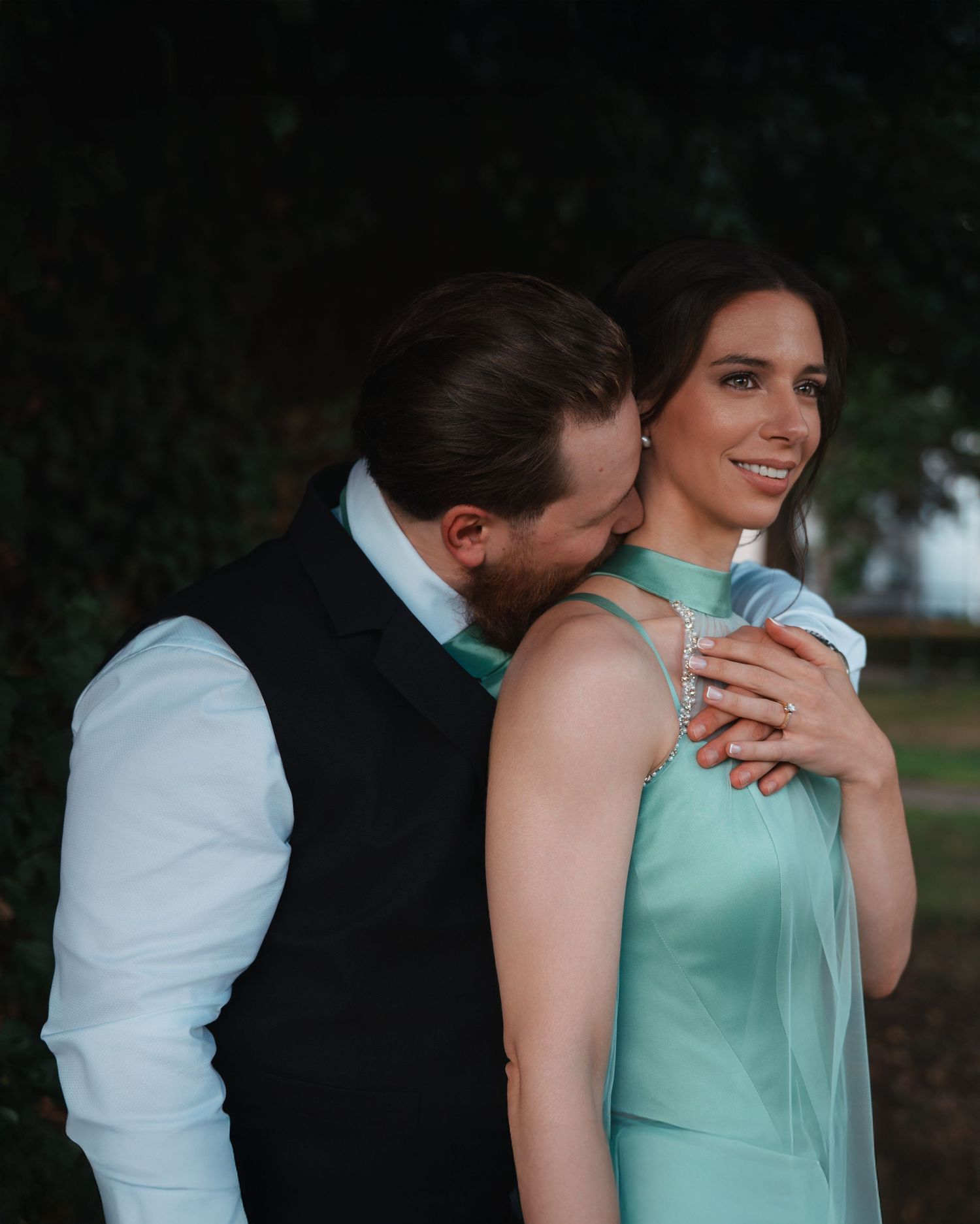 Groom in black vest kisses bride's shoulder as she smiles in a mint green dress outdoors.