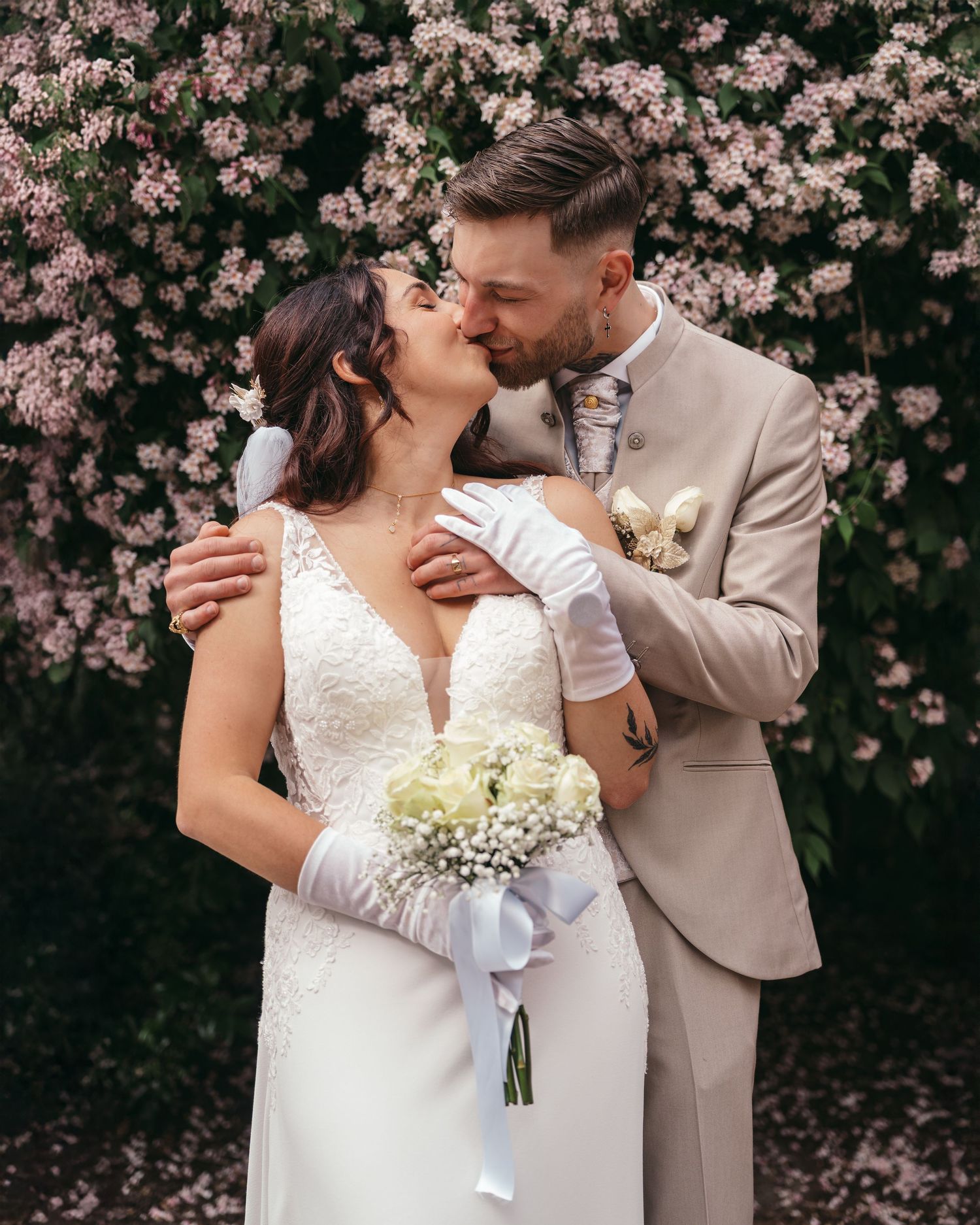 A bride and groom share a romantic kiss in front of a blooming floral backdrop, she holds white roses.