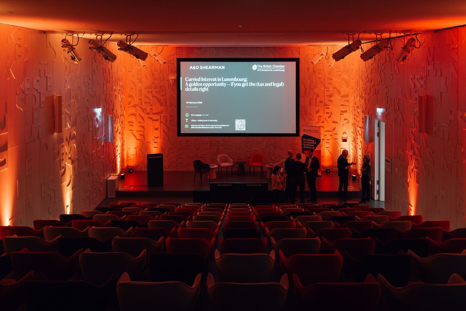 Empty cinema-style conference room with red ambient lighting and presentation screen on stage.