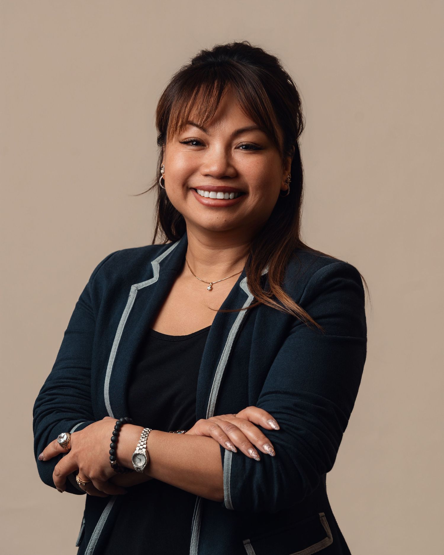 Smiling Asian woman in a navy blazer with arms crossed, wearing a watch and necklace against a neutral beige background.