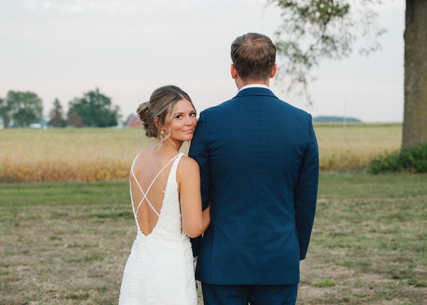 A couple in wedding attire stands together overlooking a rural field during sunset.