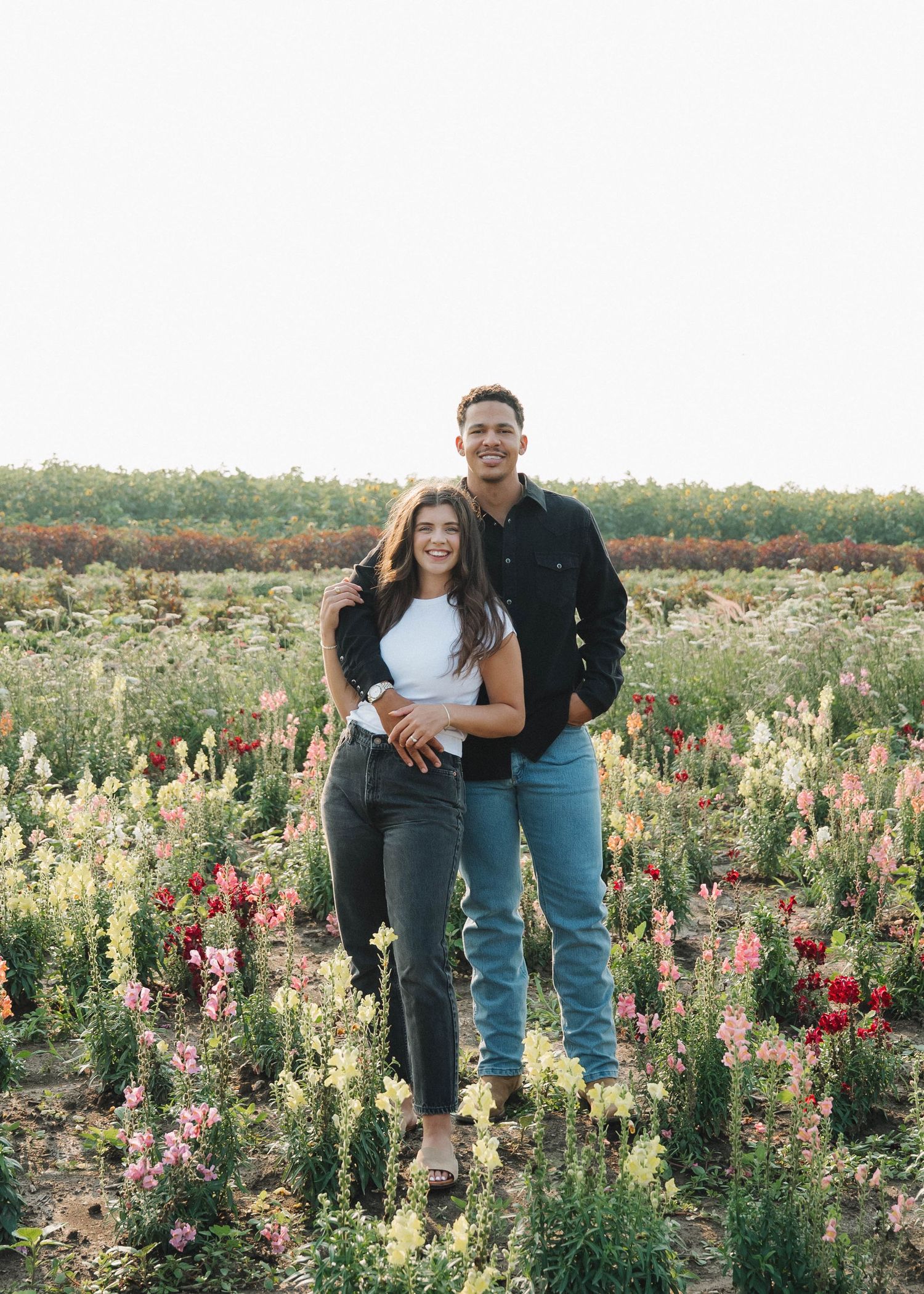 A couple stands together in a scenic field of vibrant pink and white snapdragon flowers during golden hour.