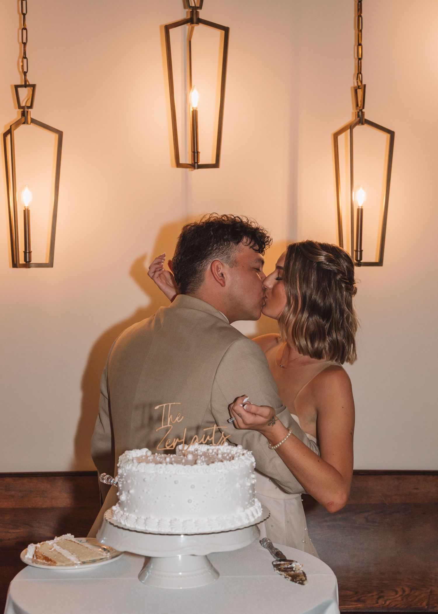 Newlyweds share cake-cutting kiss under elegant pendant lighting at reception.