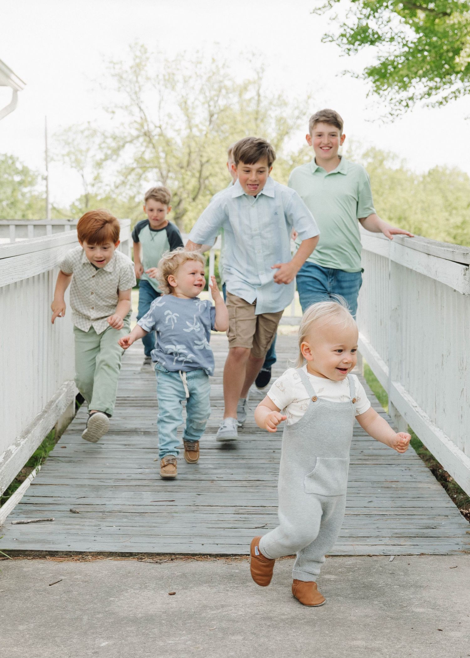 Four candid photos show playful children walking and jumping along a wooden boardwalk path surrounded by greenery.