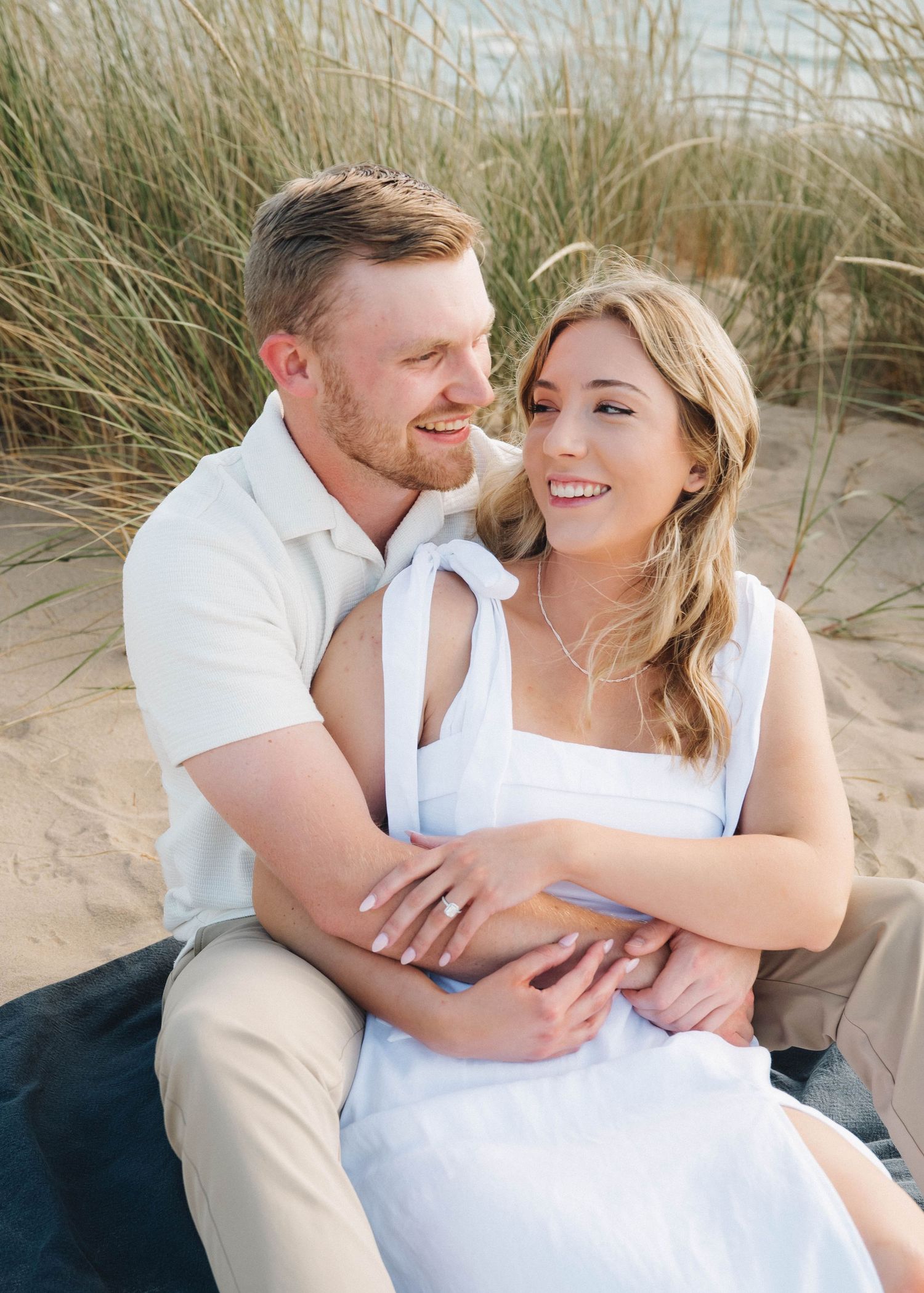 A romantic couple embraces on sandy dunes with beach grass swaying in the background at sunset.