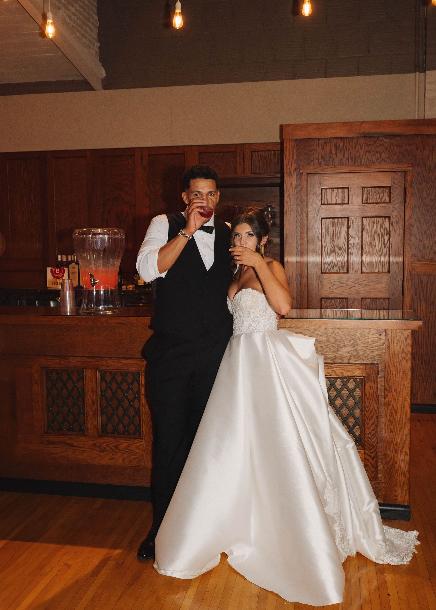 A wedding couple dances together in a wood-paneled reception hall with pendant lights hanging from above.