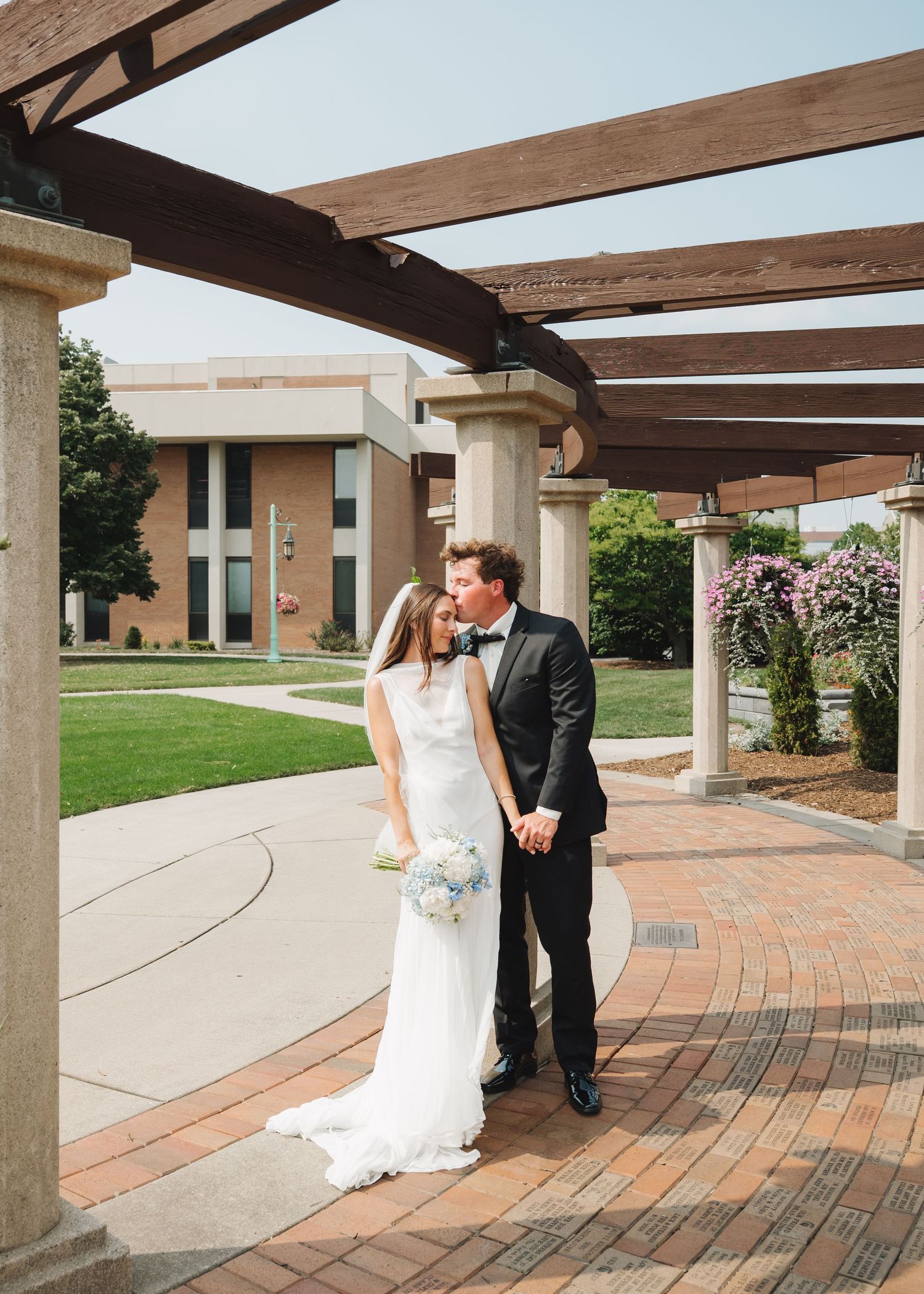 A bride and groom share a romantic kiss under a wooden pergola on a brick patio at an outdoor wedding venue.