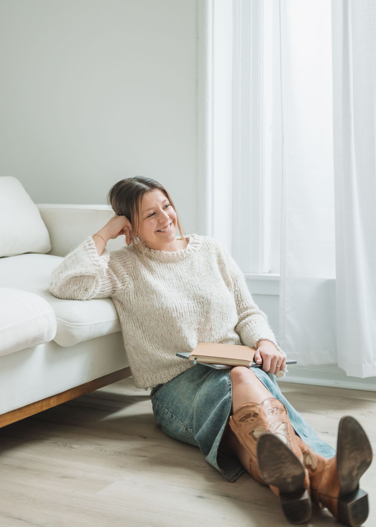 Relaxed indoor portrait of person in cream sweater reading book while sitting on floor by white couch.