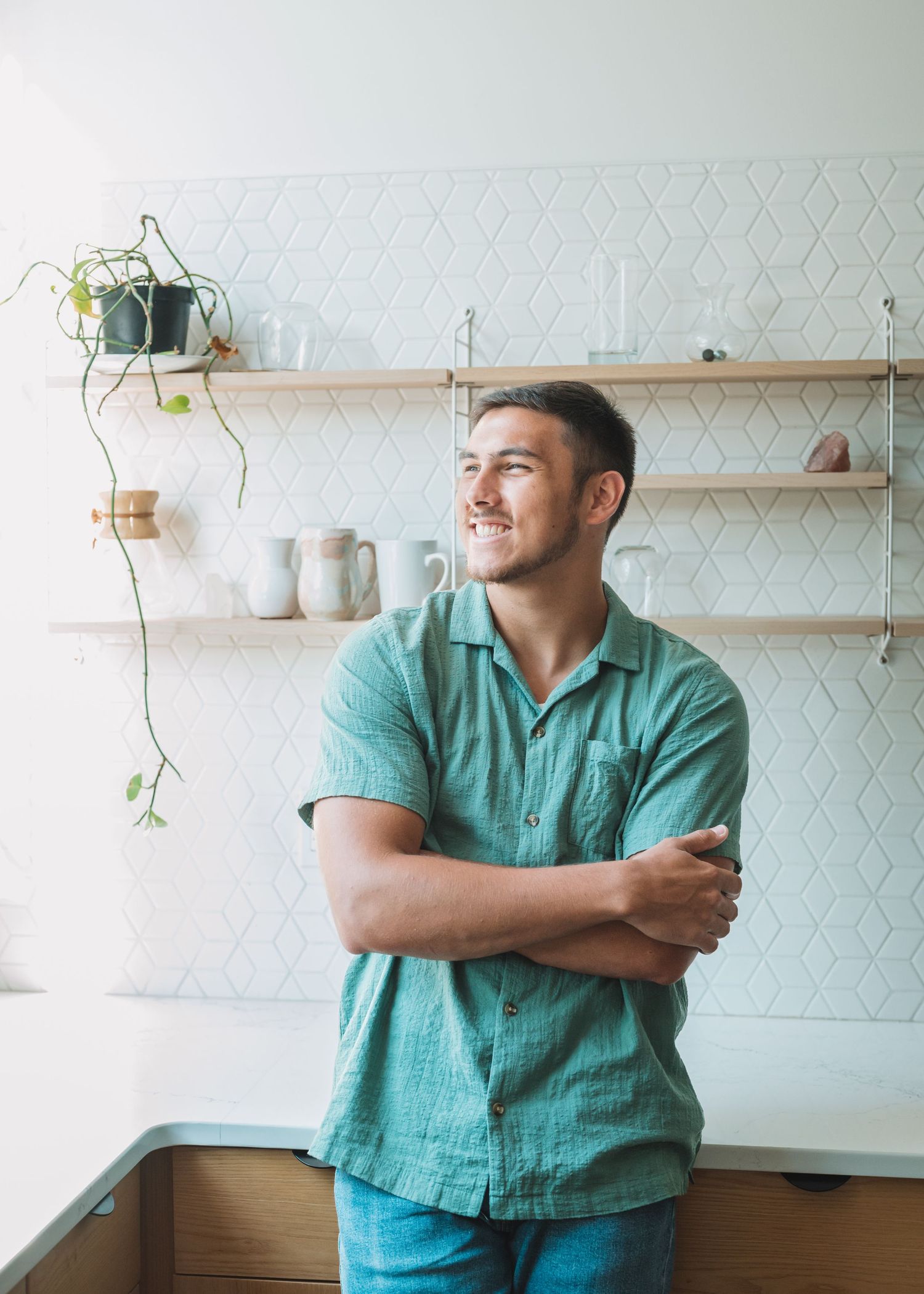 Person in teal shirt standing in modern kitchen with floating shelves and geometric backsplash.