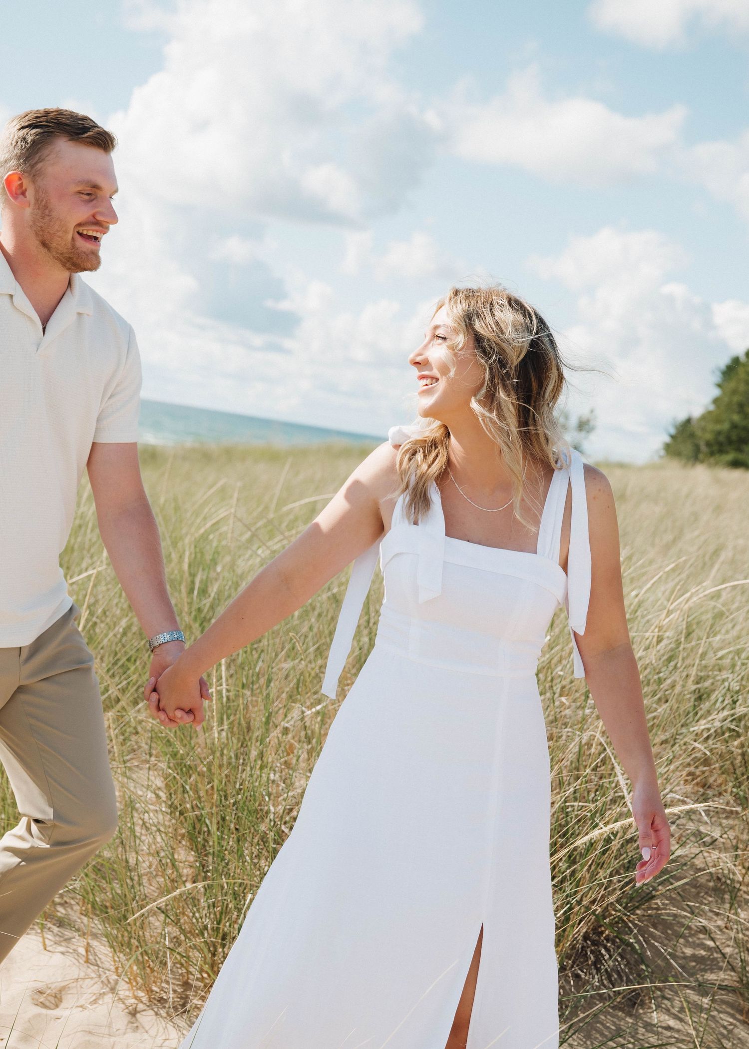 A couple walks joyfully through tall beach grass on a sunny day, with the woman wearing a white dress.