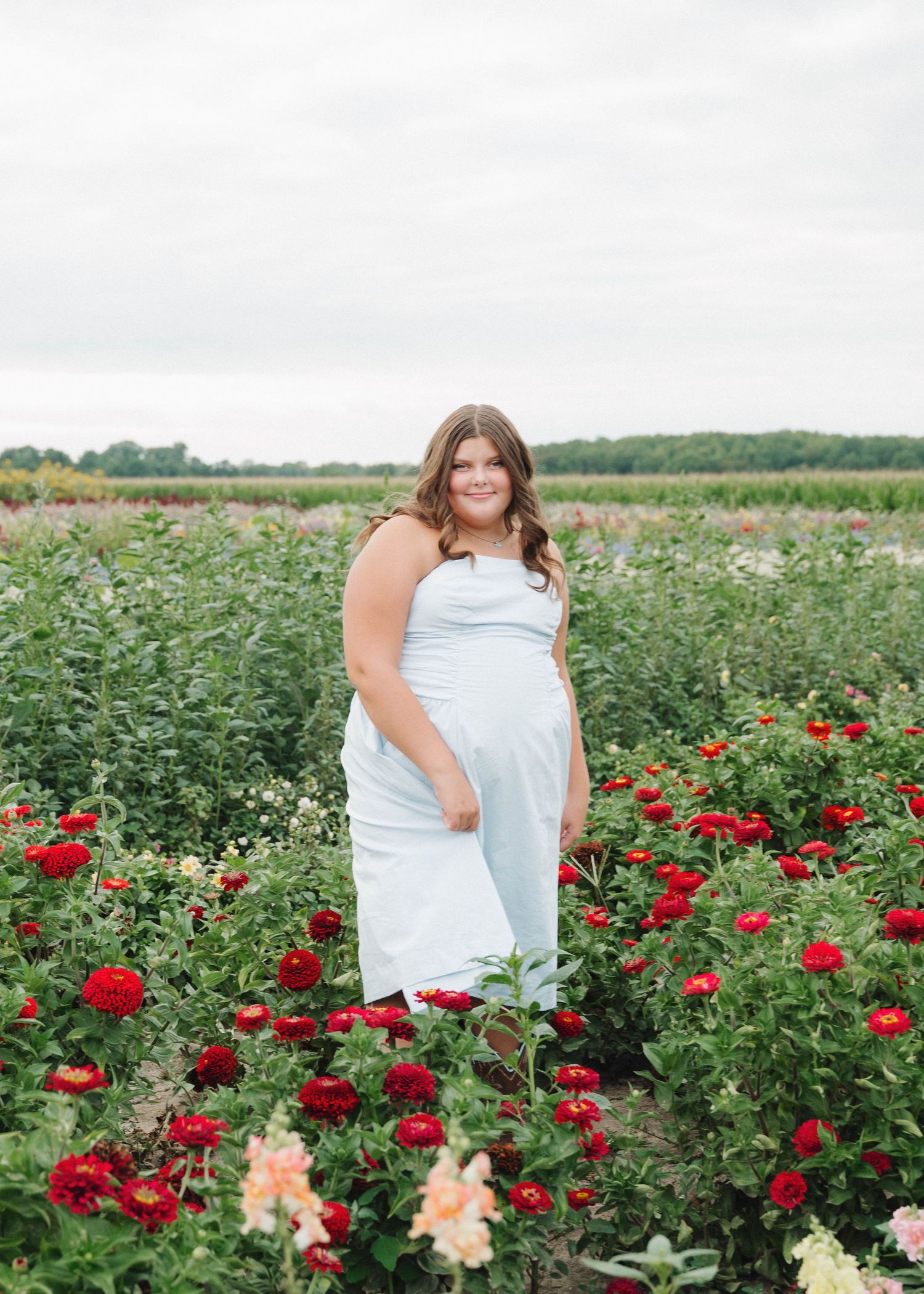 A person in a white dress stands in a garden filled with blooming red and peach-colored roses on a sunny summer day.