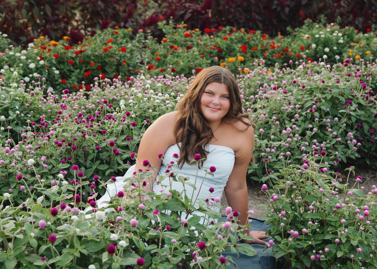 Someone in a white dress poses among colorful blooming flowers in a lush garden setting.