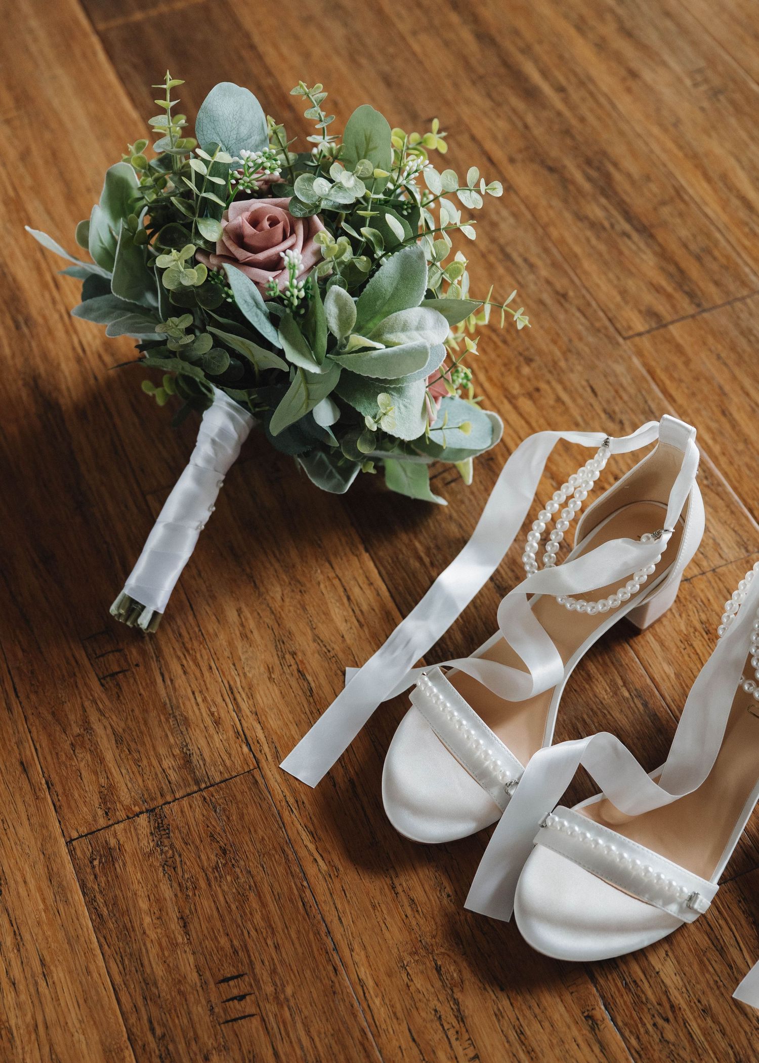 Elegant bridal bouquet with succulents and mauve roses next to white strappy wedding sandals on wooden floor.