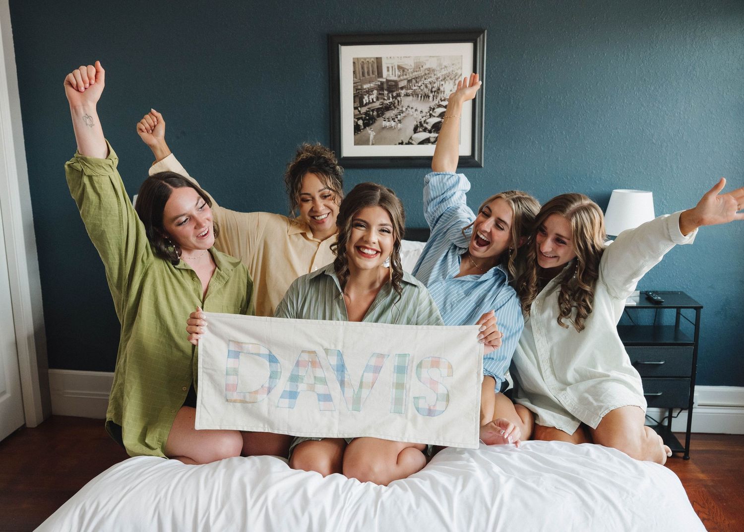 A group celebrates joyfully on a bed while holding up a sign together in a bedroom with teal walls.
