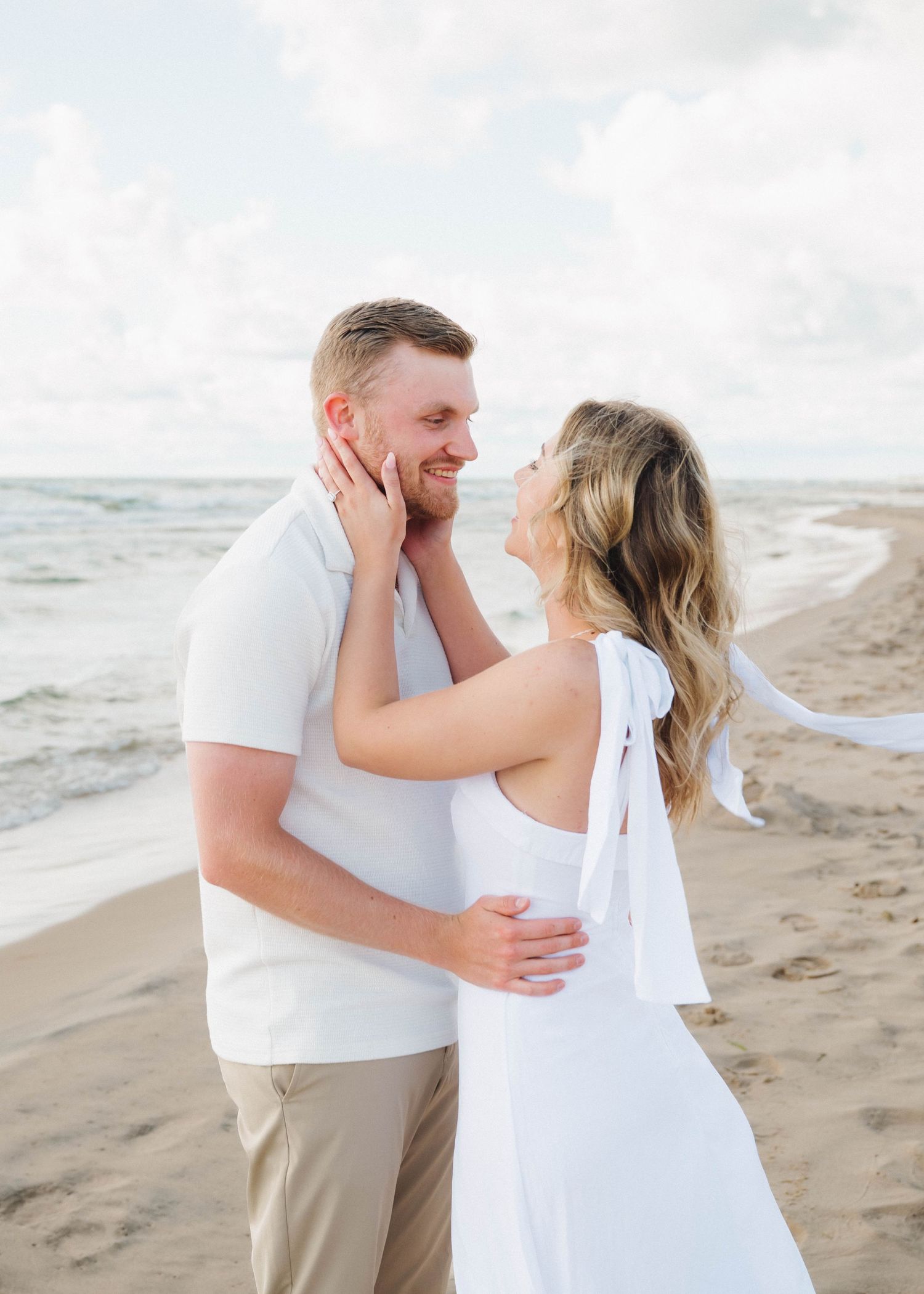 A couple in white attire shares a romantic moment on a sandy beach with ocean waves in the background.