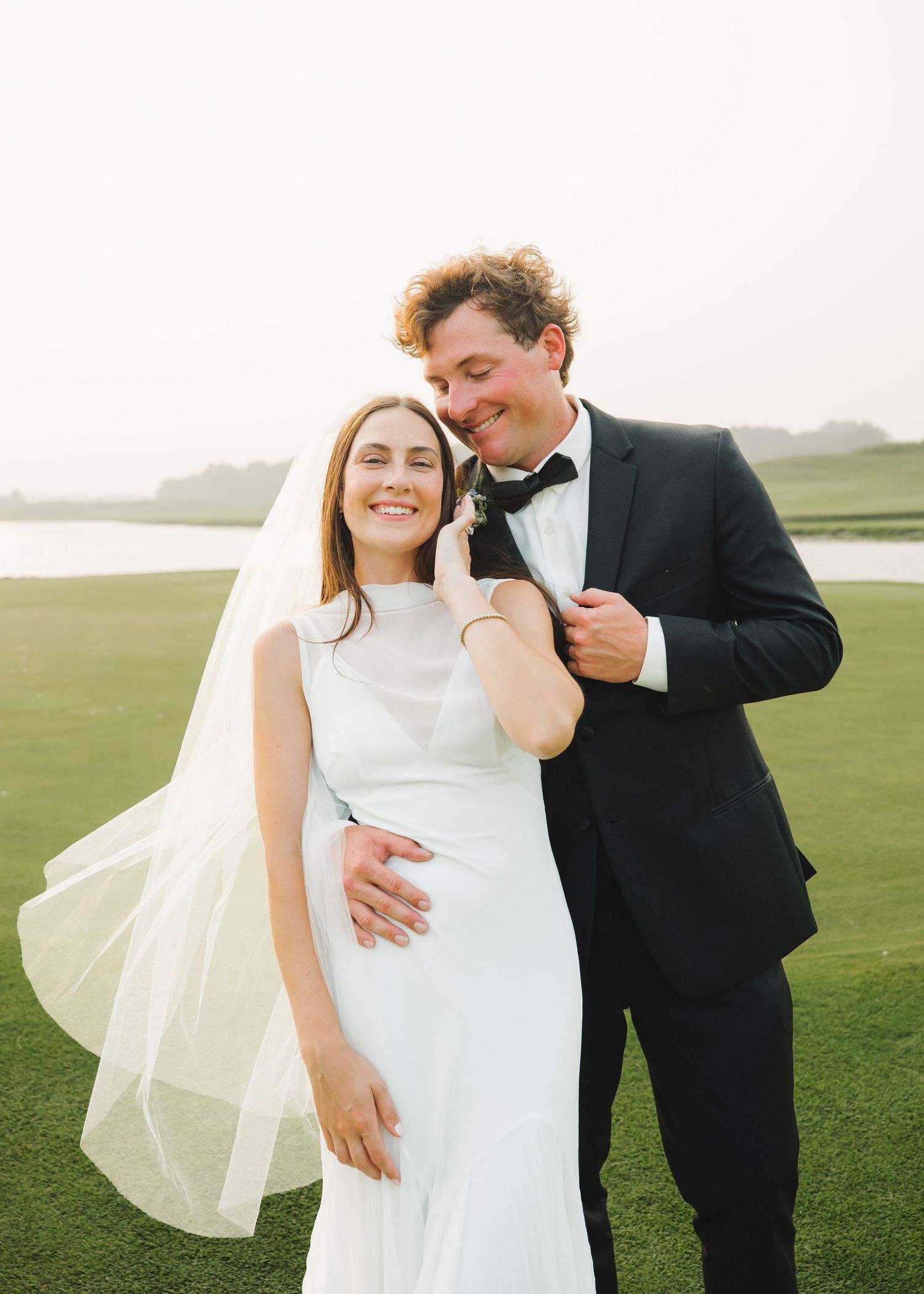 Bride in white gown and veil smiles as groom in black tuxedo embraces her on a green lawn.