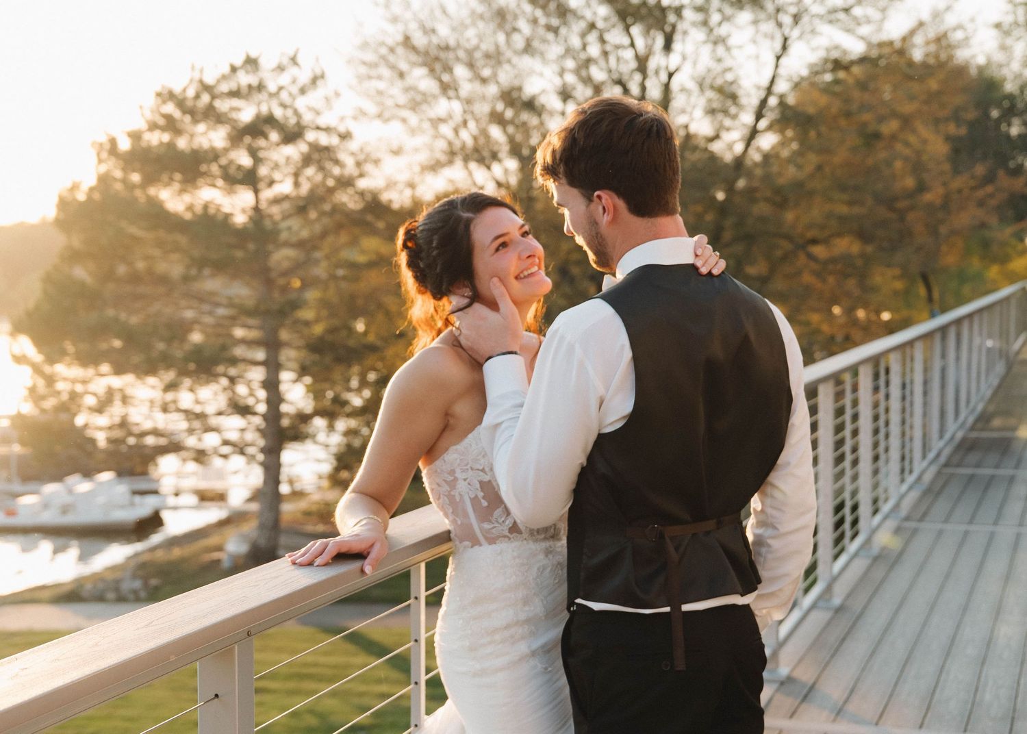 A couple embraces on a waterfront deck during golden hour sunset as trees sway in the background.