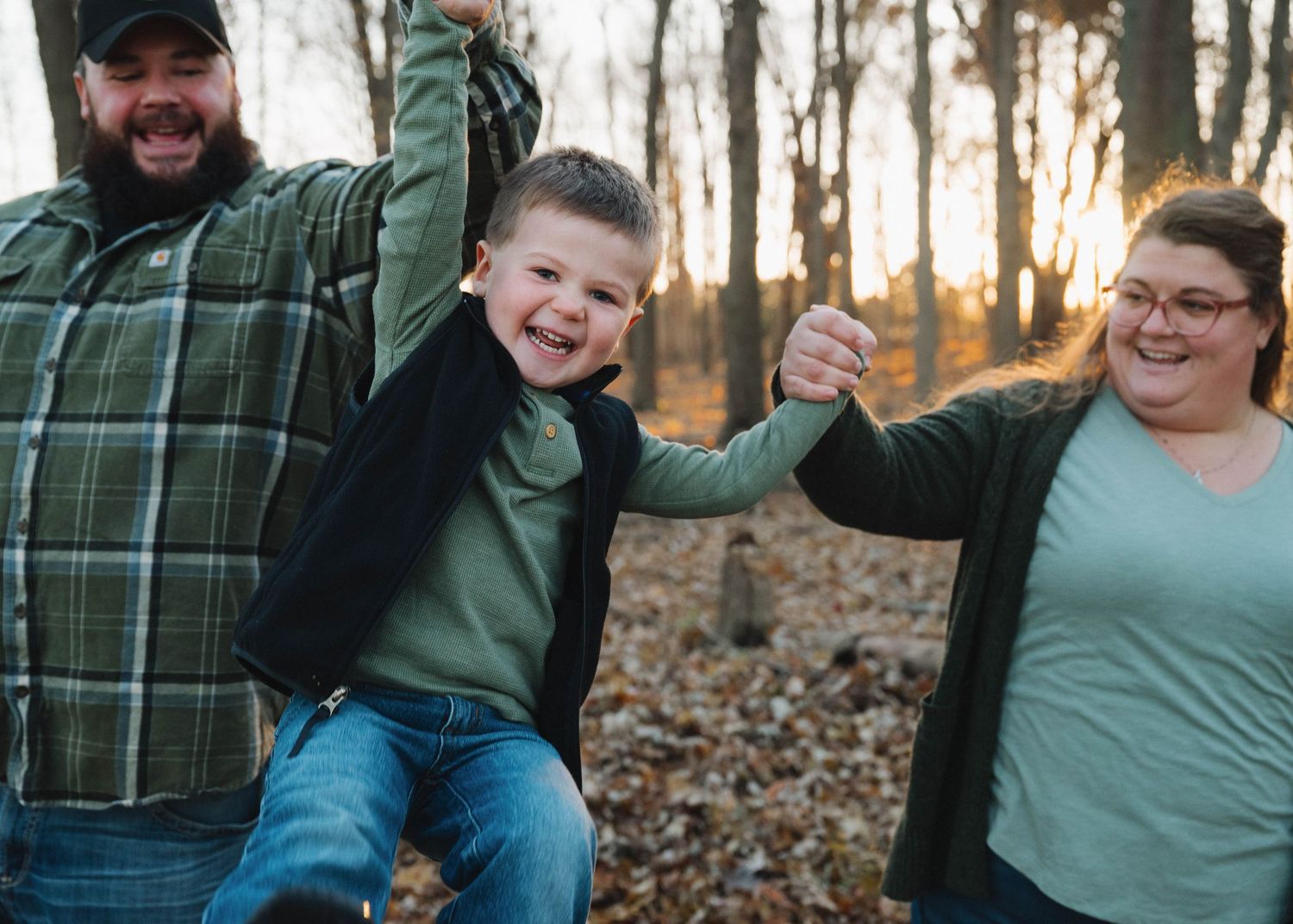 A family in matching green outfits swings a child between them while walking through autumn woods at sunset.