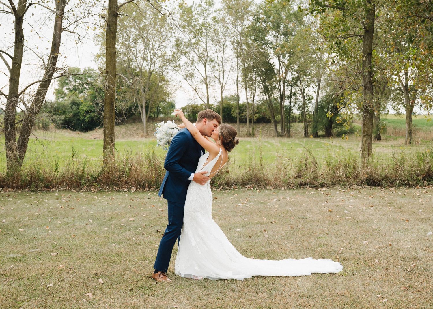 A couple in wedding attire embraces in a rustic outdoor setting with trees and natural scenery in the background.