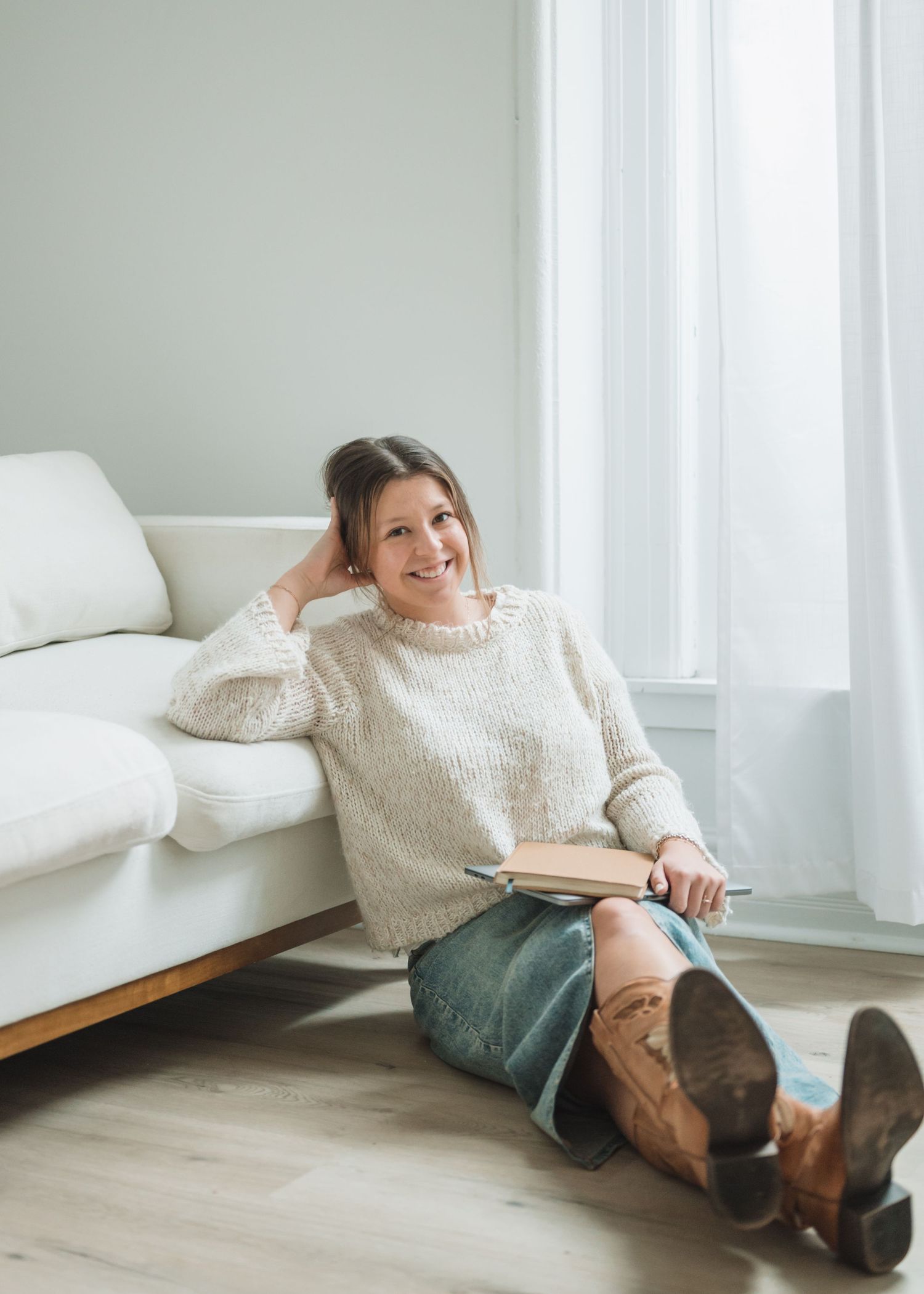Person relaxing on floor wearing cozy cream sweater and boots next to bed in bright minimalist room.