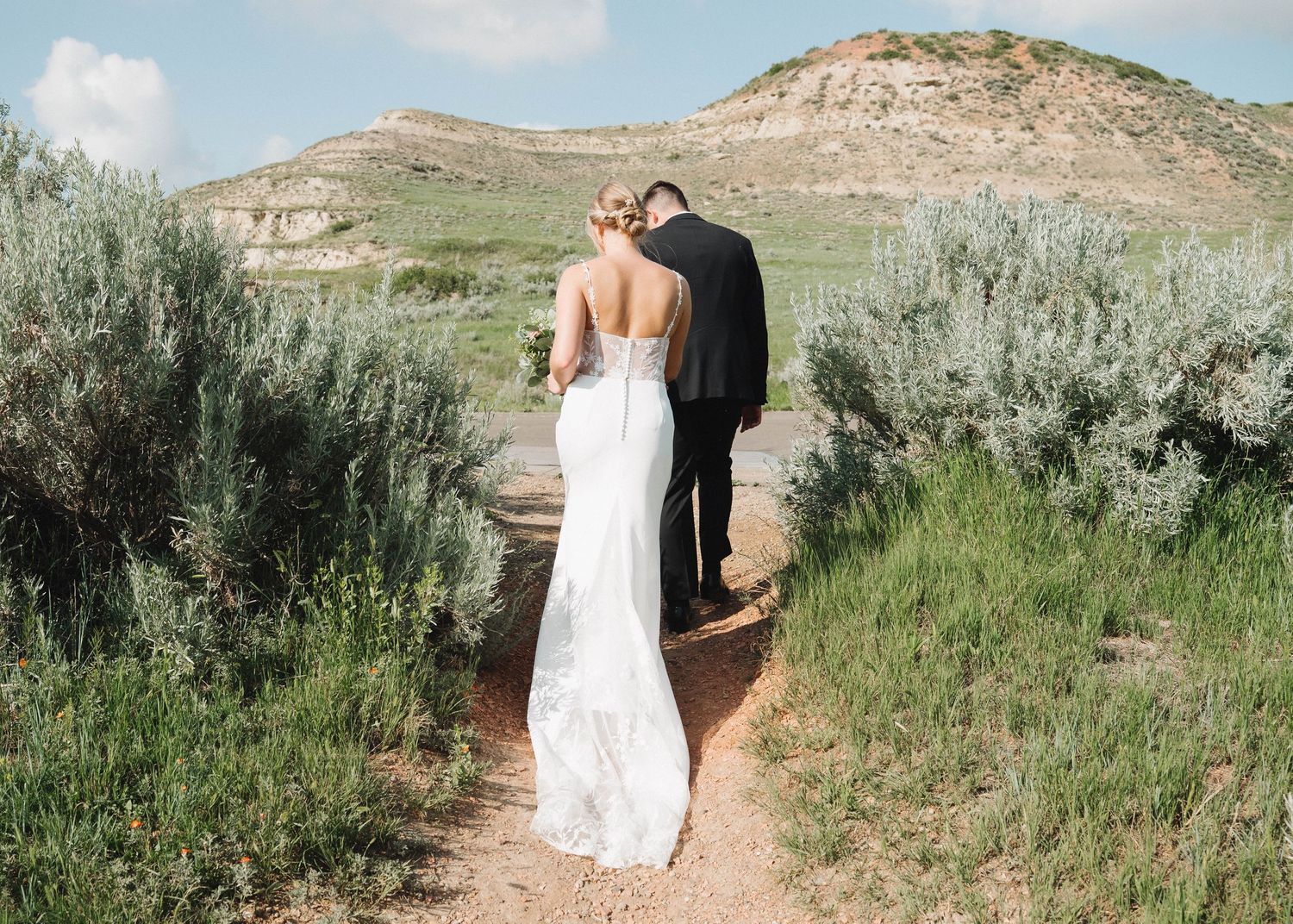 A person in a white wedding dress walks along a winding dirt path through desert sage brush and mountains.