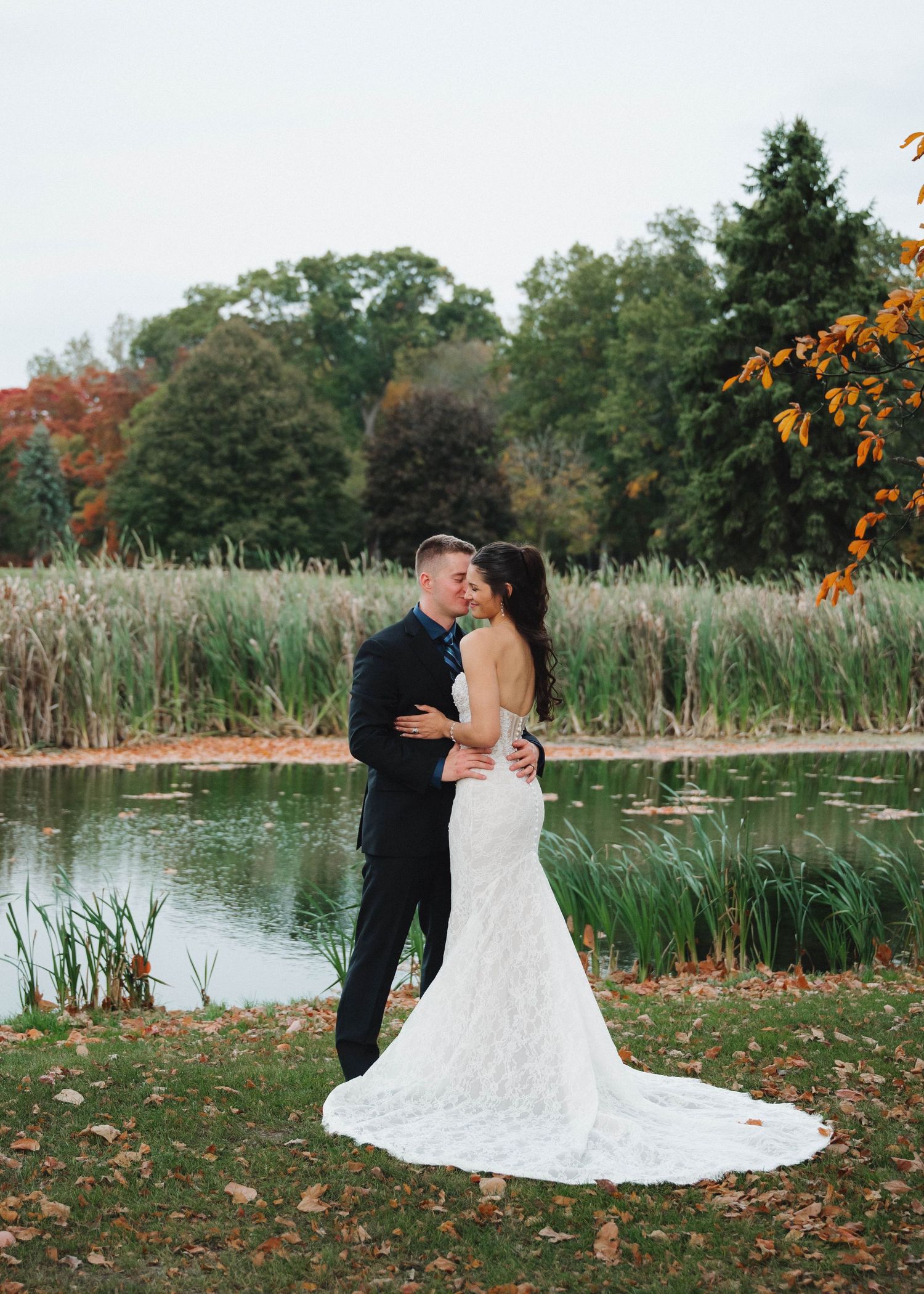 A couple shares a romantic moment by a pond surrounded by autumn foliage and cattails.
