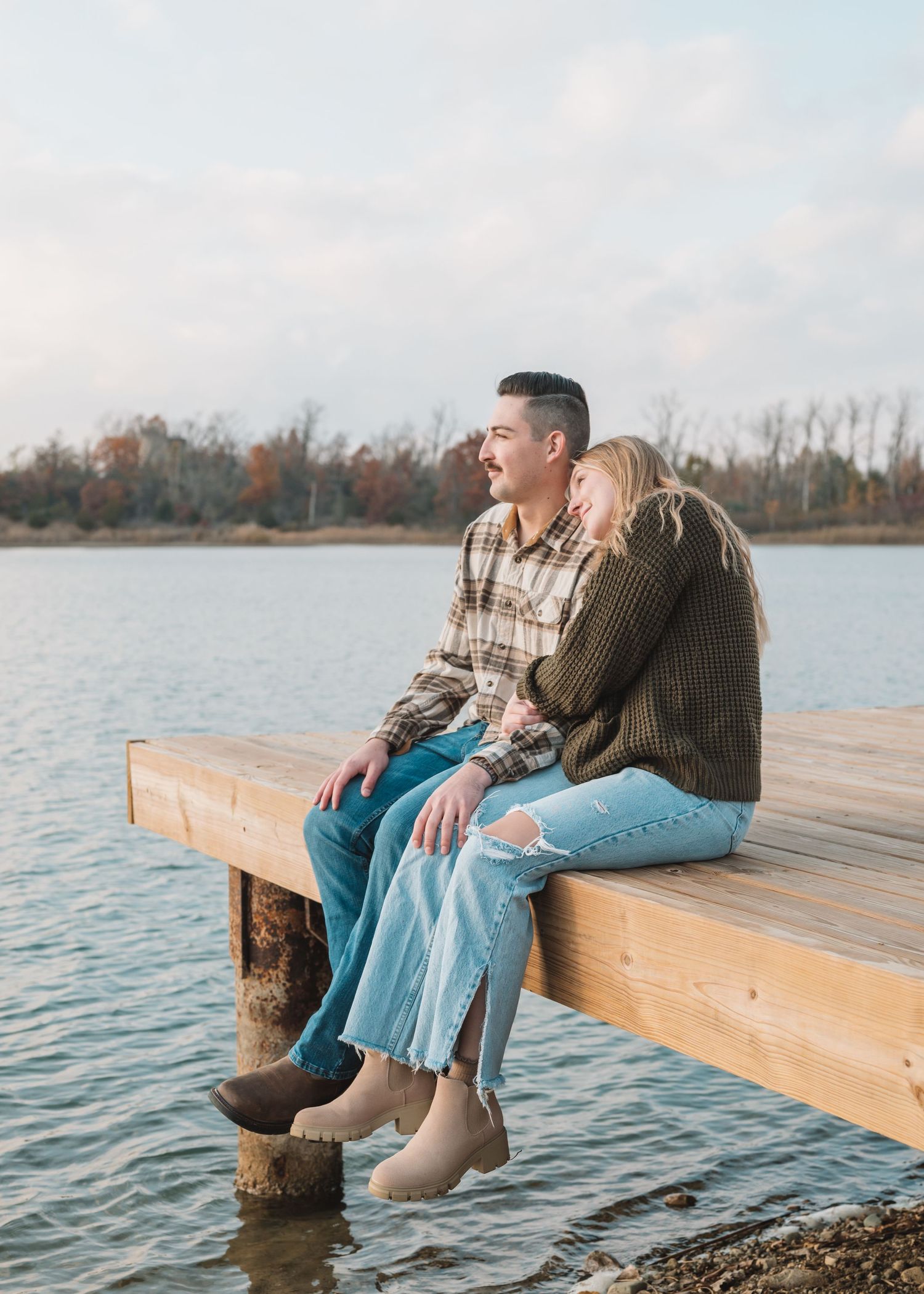 A romantic moment captured on a lakeside dock surrounded by autumn trees and calm waters.