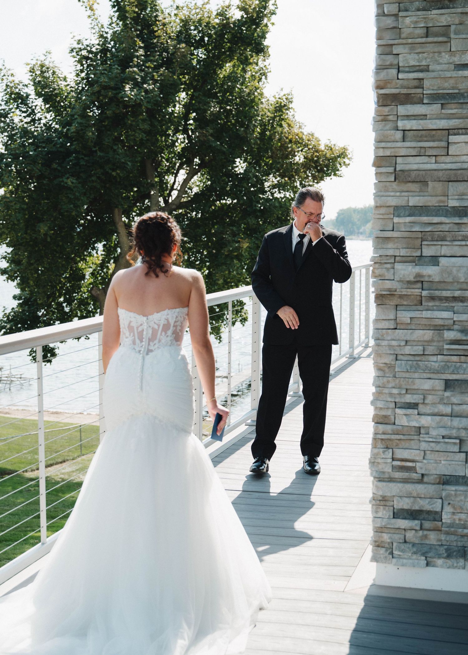 A couple shares an emotional first look moment on a lakeside balcony with stone pillars and greenery.