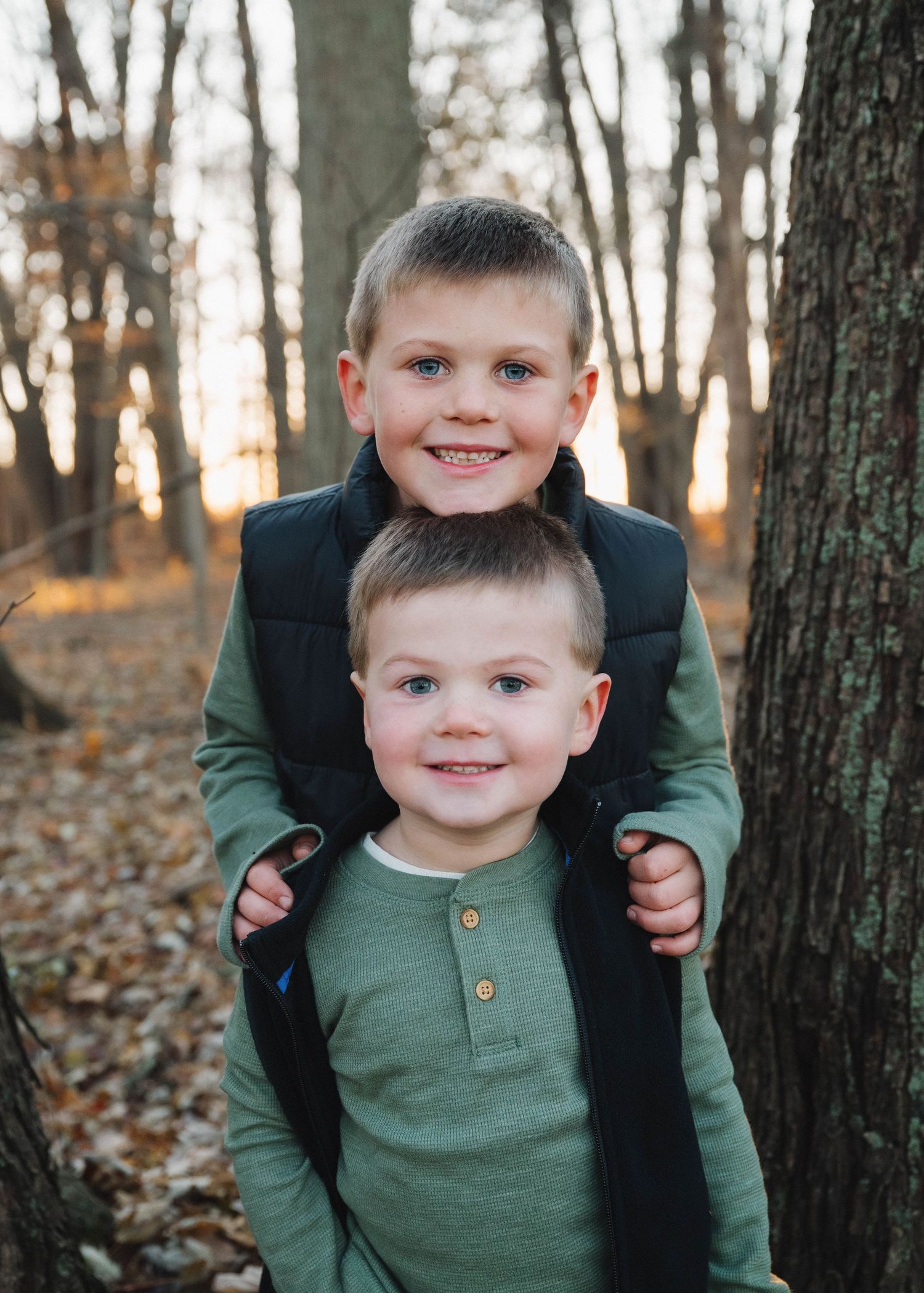 Two children in matching green shirts play among autumn trees and fallen leaves in a wooded setting.