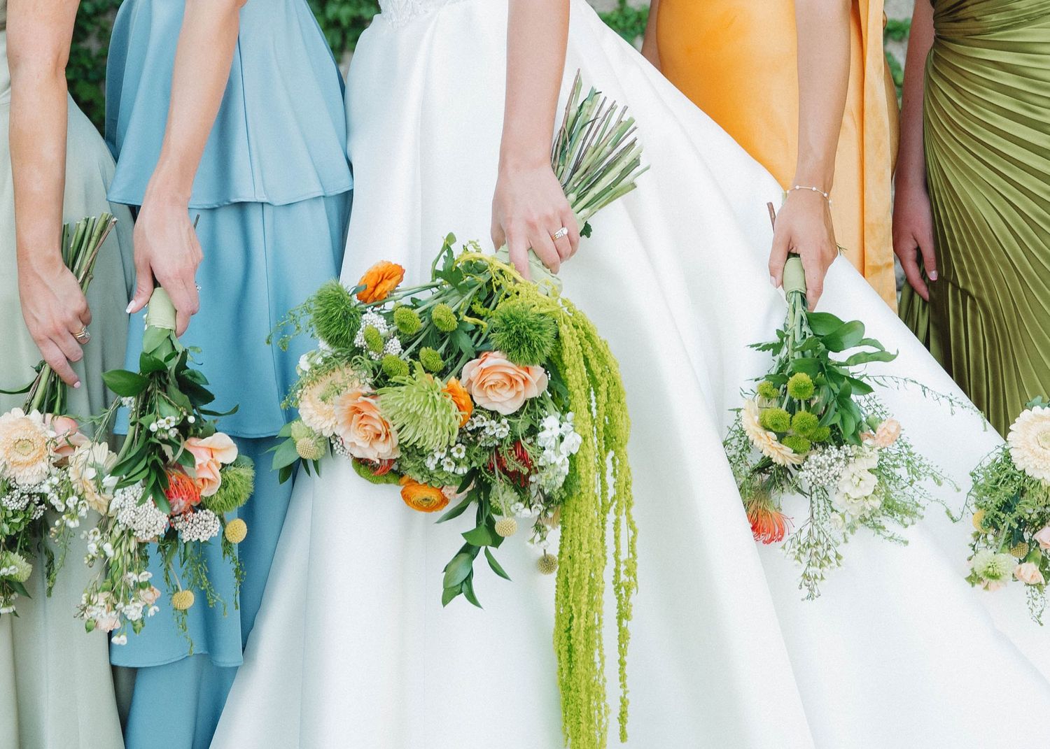 Wedding party members in pastel blue and green dresses hold vibrant bouquets with cascading greenery and peach flowers.