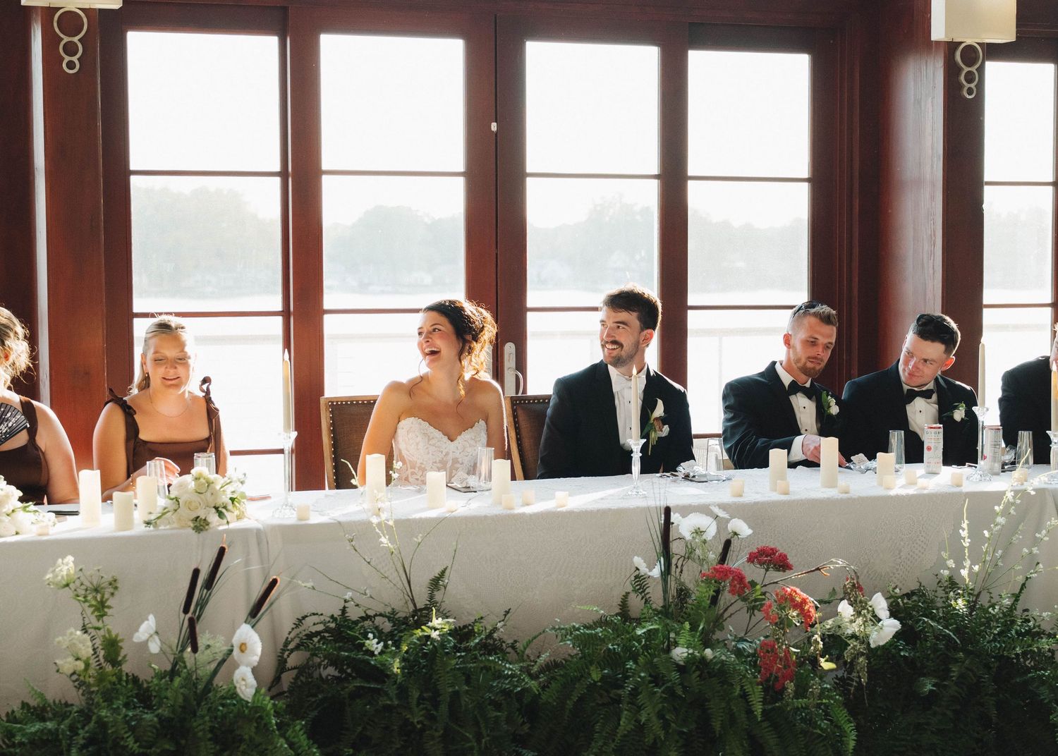 Wedding reception setup in rustic venue with large windows, long tables, and natural greenery decorations.