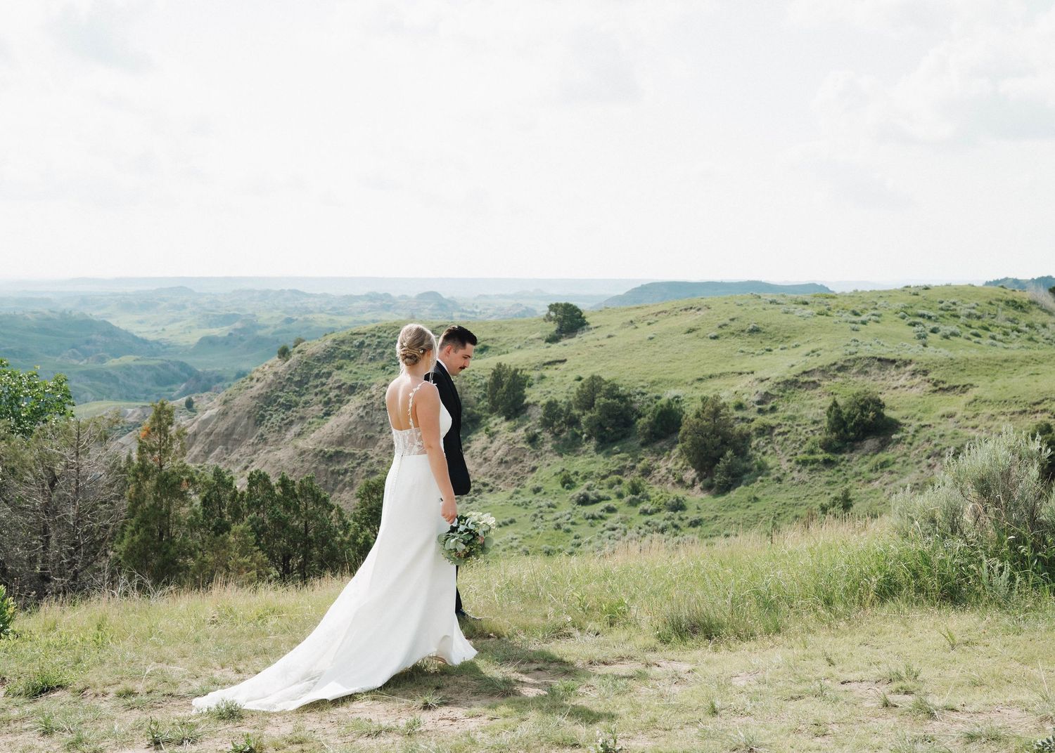 A bride and groom embrace on a scenic mountain hillside overlooking a vast green landscape on their wedding day.