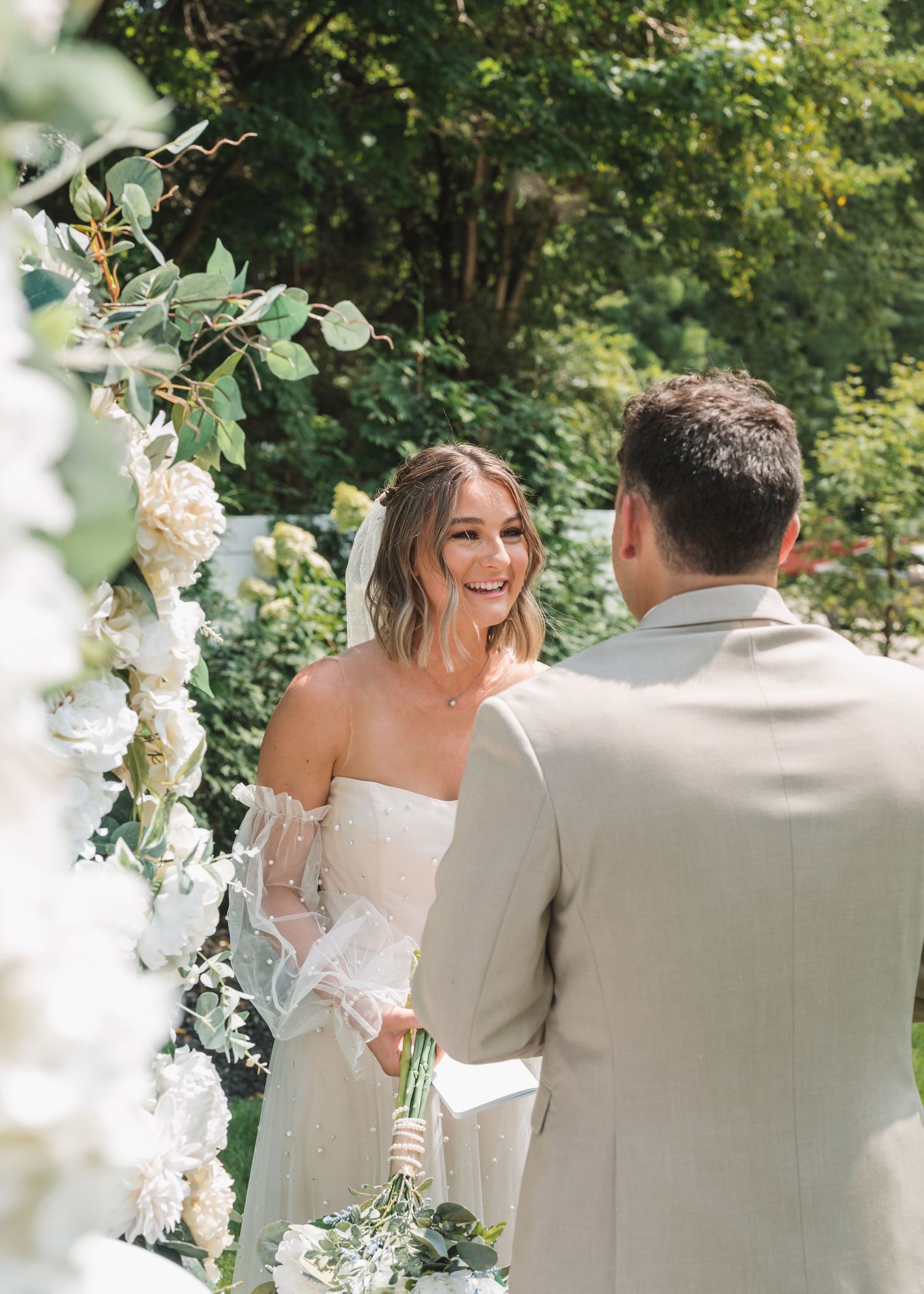 Outdoor wedding scene with white floral arch and natural greenery backdrop.