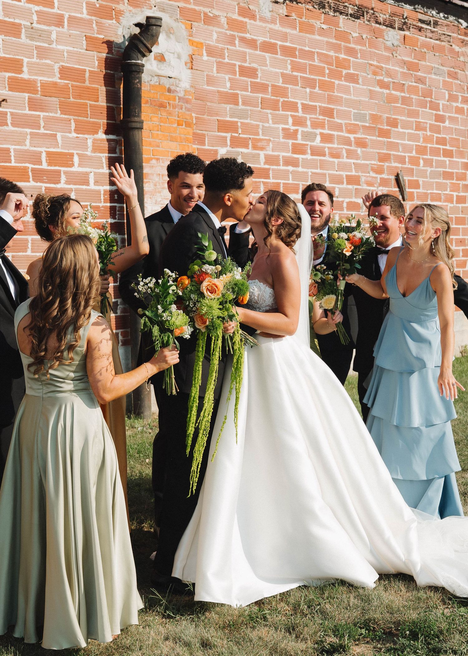 Wedding party poses against brick wall with bridesmaids in light blue dresses holding green bouquets.