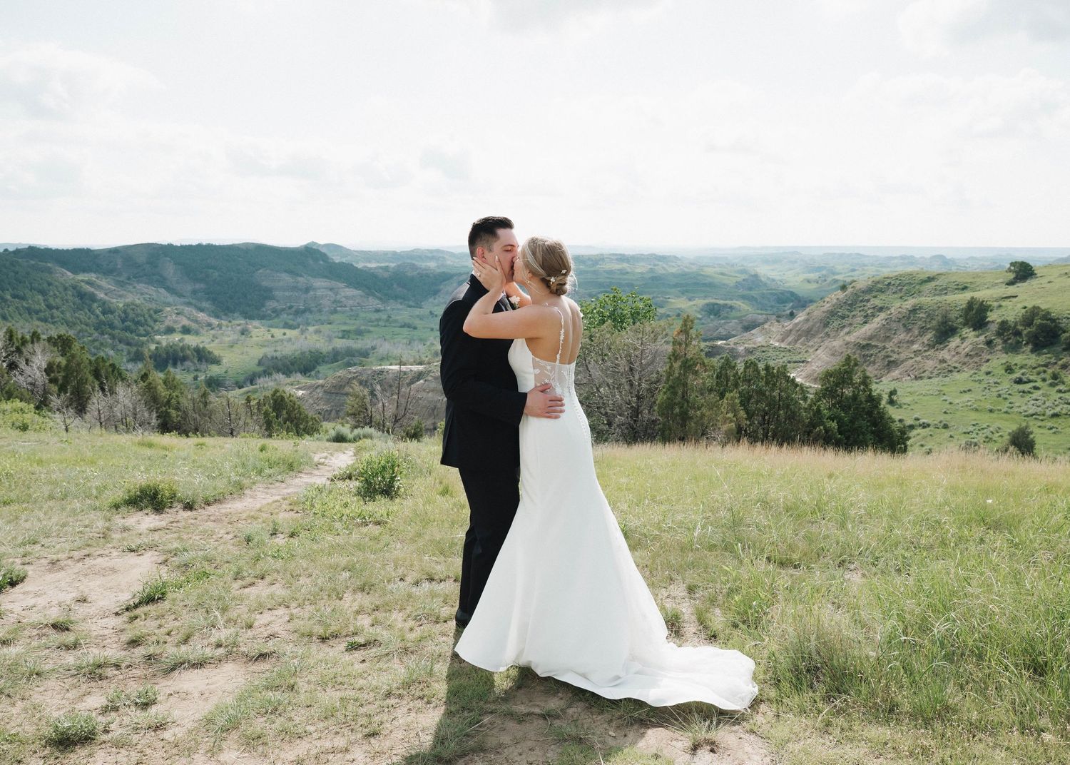A bride and groom embrace on a scenic hilltop overlooking rolling green hills and valleys.