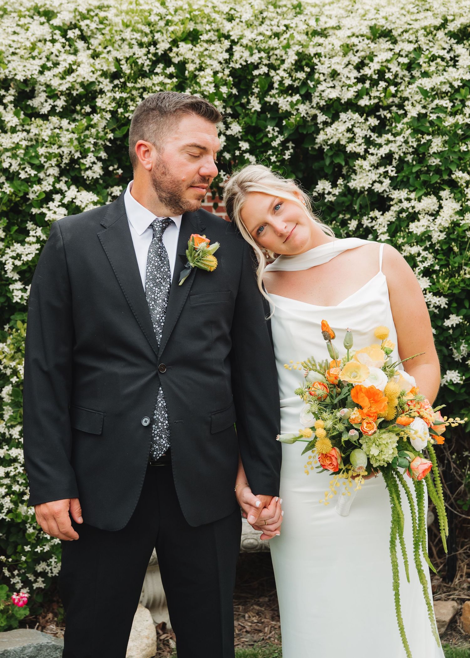 A bride and groom hold hands and smile in front of white blooming flowers, she holds an orange and green bouquet.