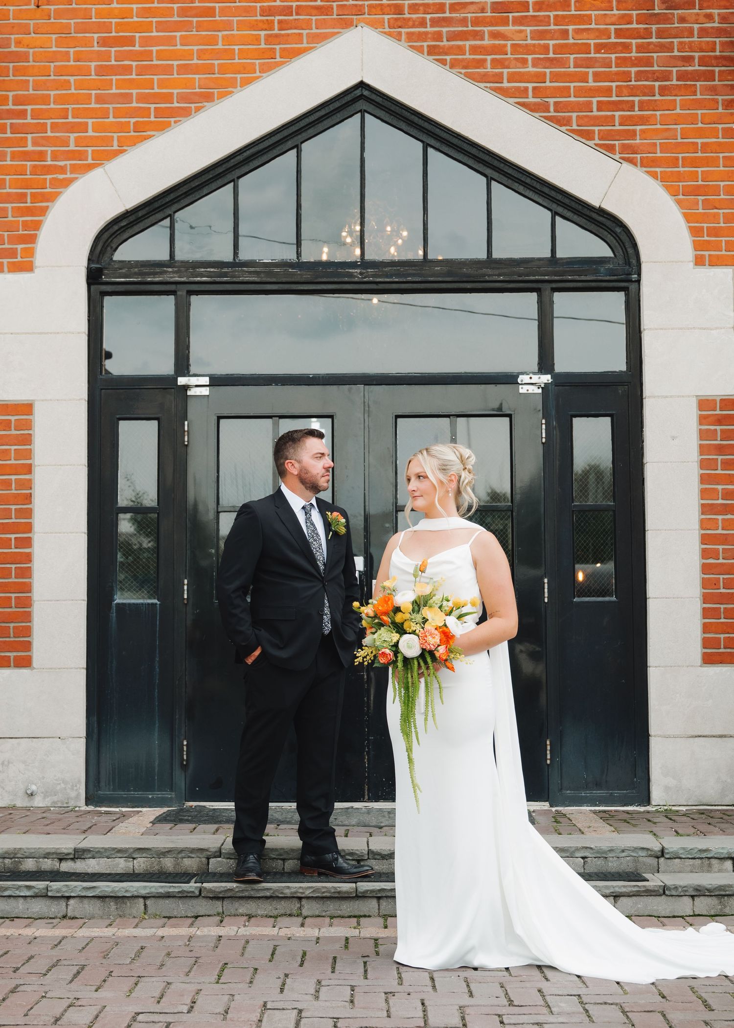 Bride and groom posing in front of brick building with arched glass doors, bride holding vibrant orange bouquet.