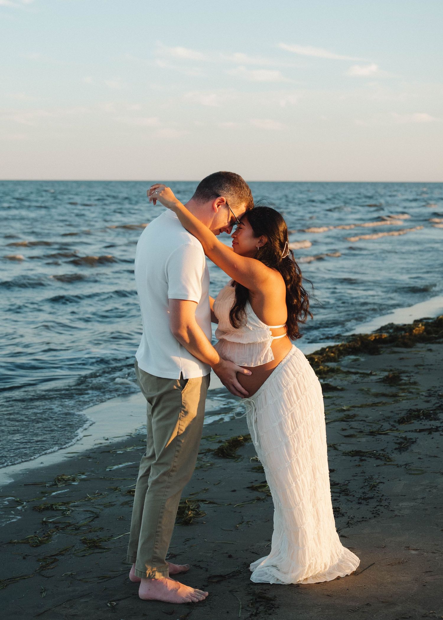 A couple shares a romantic kiss on a serene beach at sunset with gentle waves in the background.